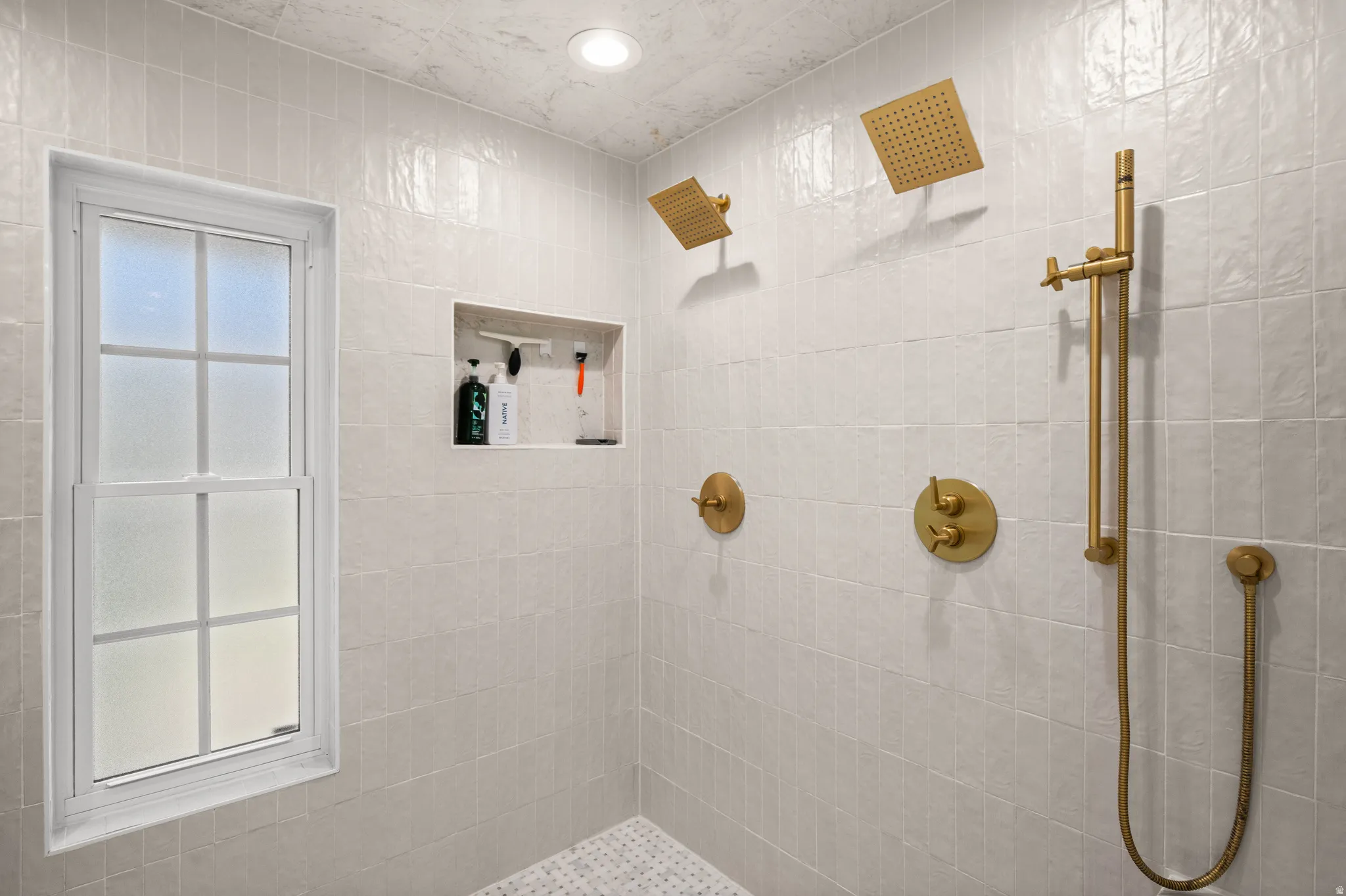 Shower featuring gold-finish fixtures, dual square rain showerheads, a hand-held sprayer, and a built-in tiled niche