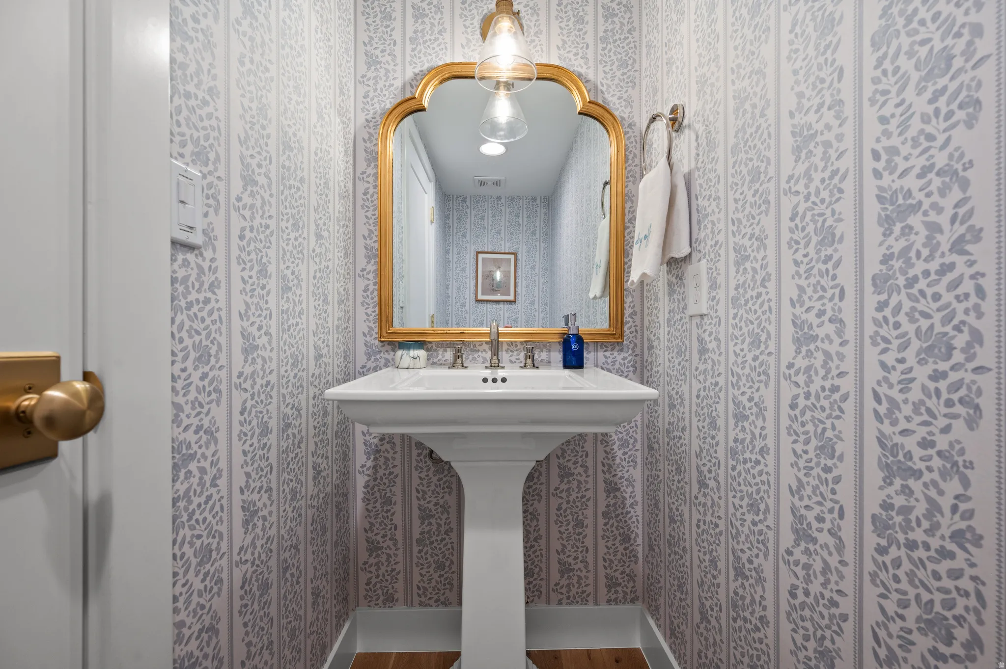 Pedestal sink with brass-finish faucet, framed by a gold-tone mirror and patterned wallpaper