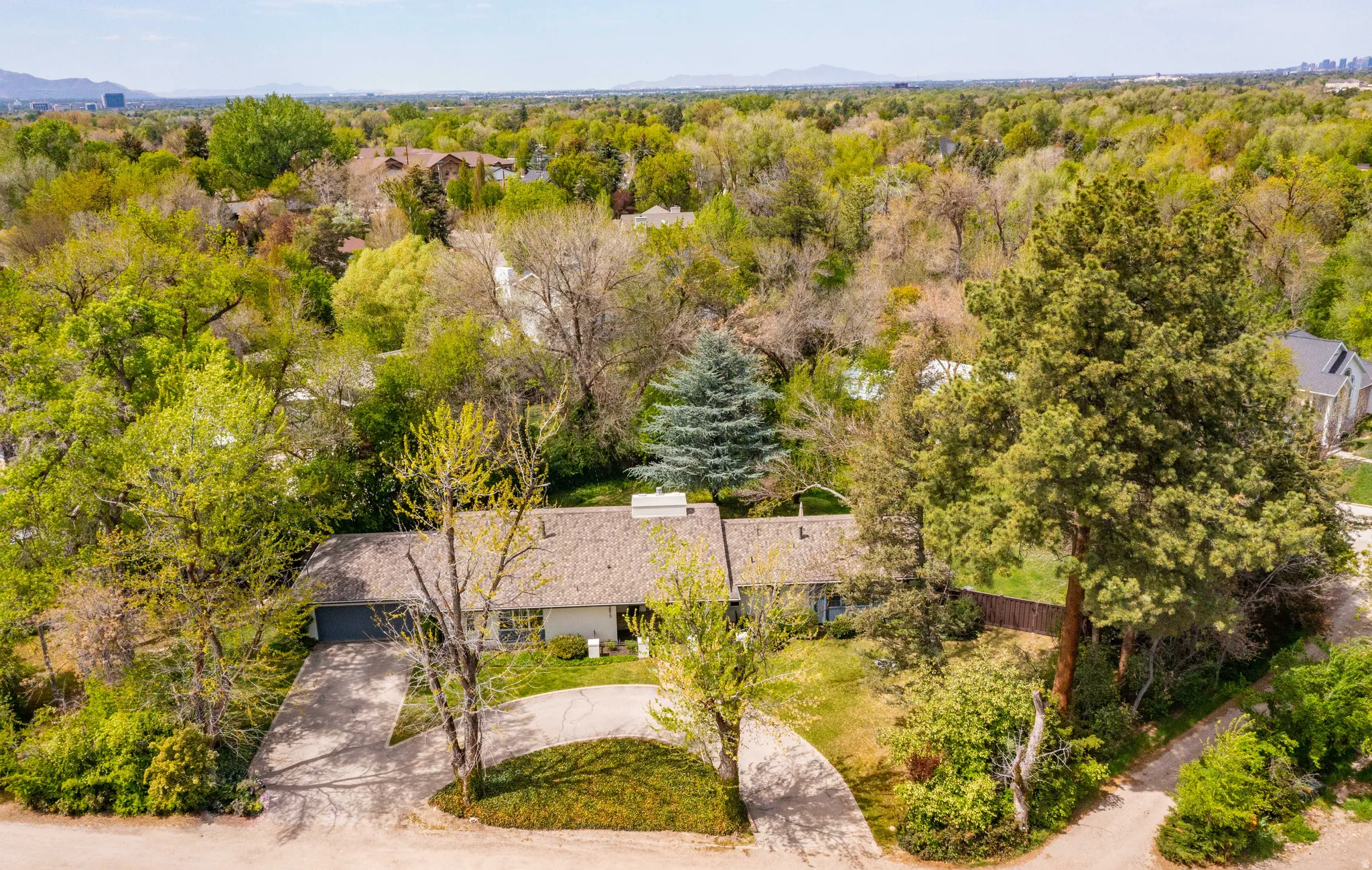 Ranch-style residence featuring a prominent front driveway with a circular turnaround, a shingled roof, a mature tree canopy, and a verdant surrounding landscape