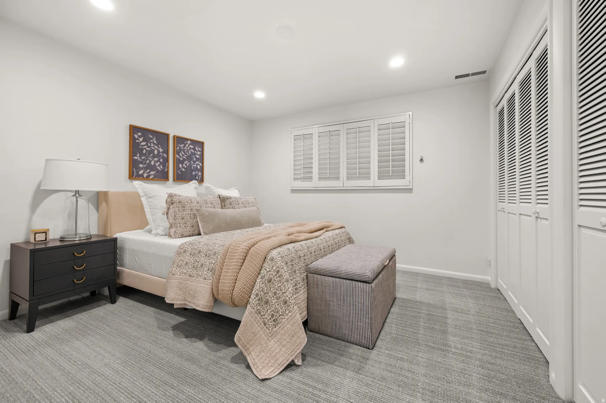 Light gray carpeted flooring, recessed ceiling lighting, a window with white plantation shutters, and louvered bifold closet doors