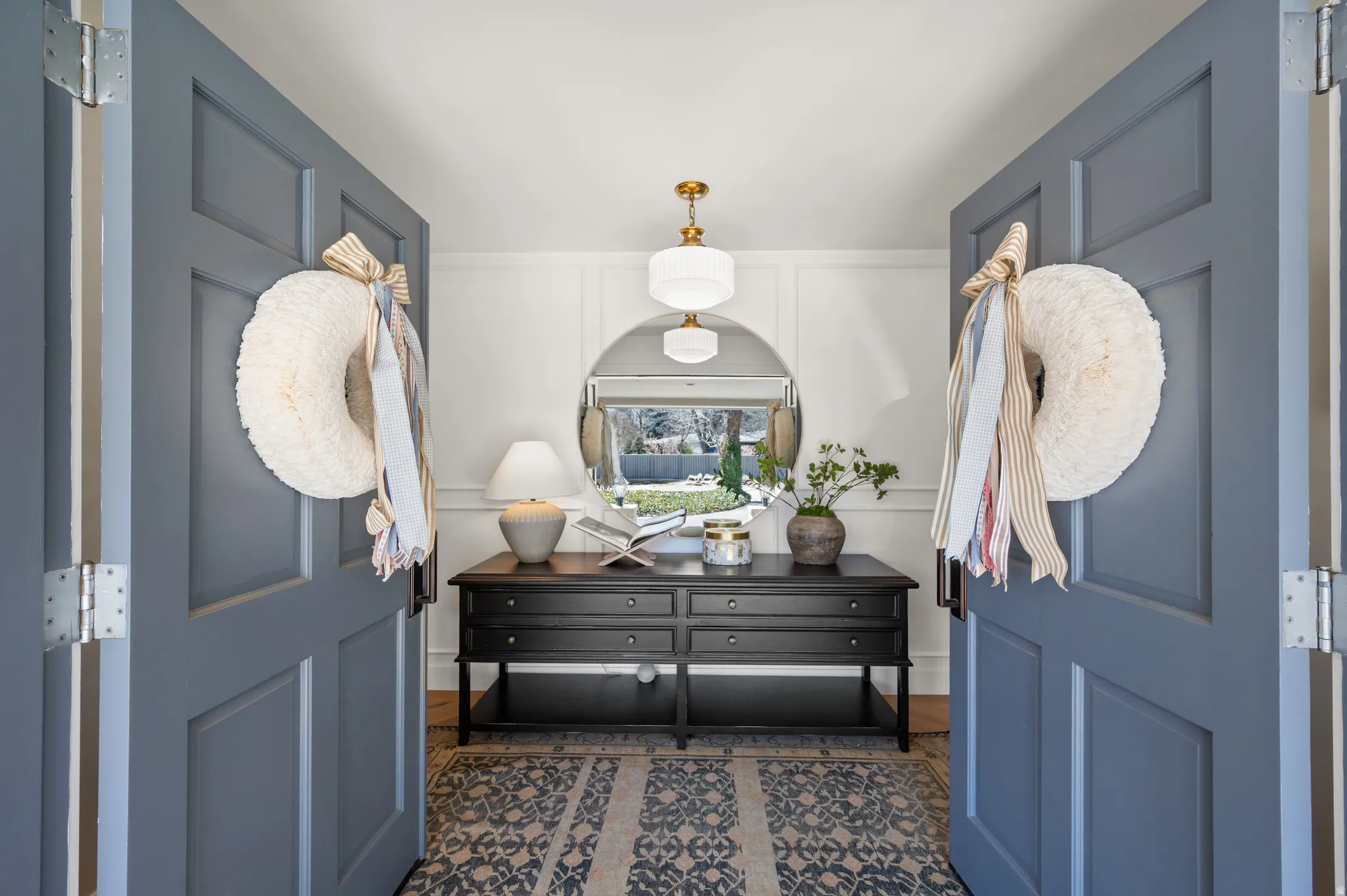 Double panel doors opening to an entryway featuring a dark wood console table, a large circular mirror, and a multi-tiered pendant light