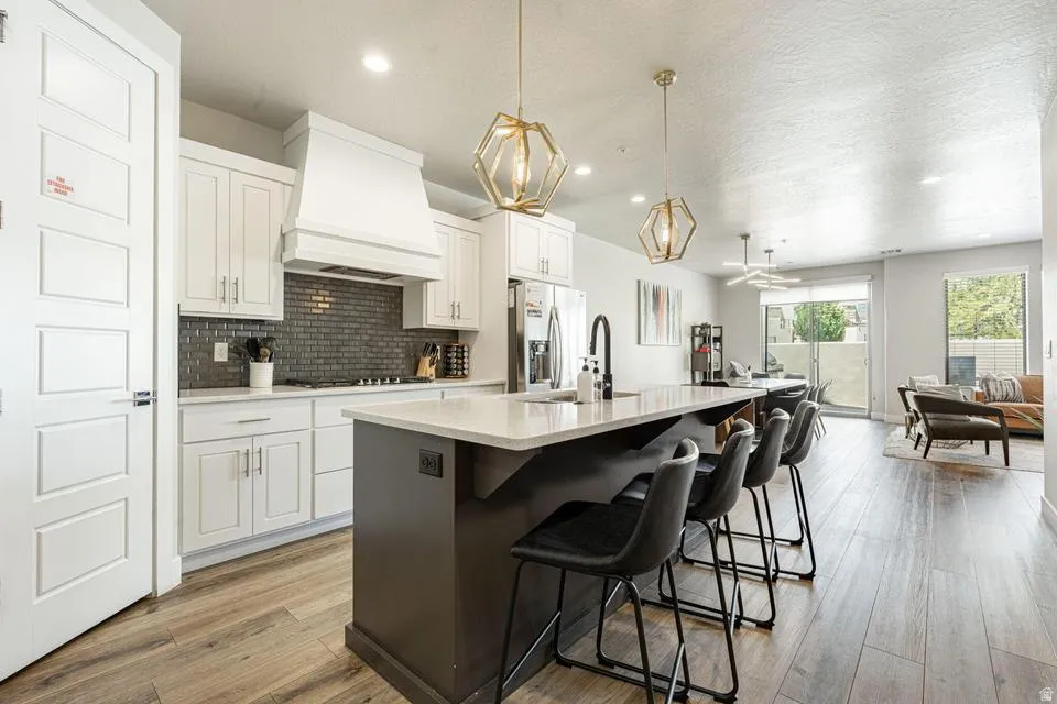 Modern kitchen featuring a dark-toned island with seating, pendant lighting, white cabinetry, a tiled backsplash, and wood-finish flooring