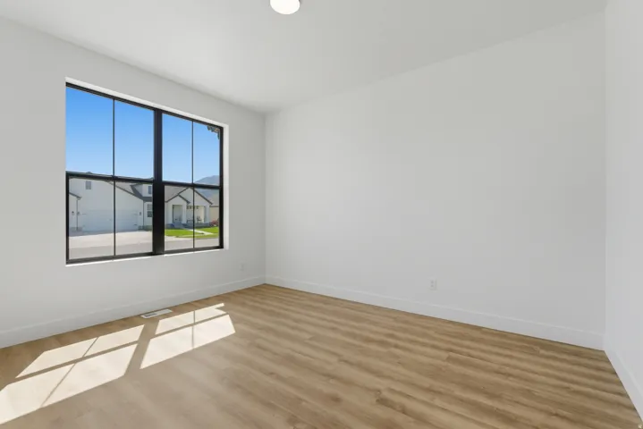Bright interior room featuring a large black-framed picture window, light wood-finish flooring, white baseboards, and a flush-mount ceiling light fixture