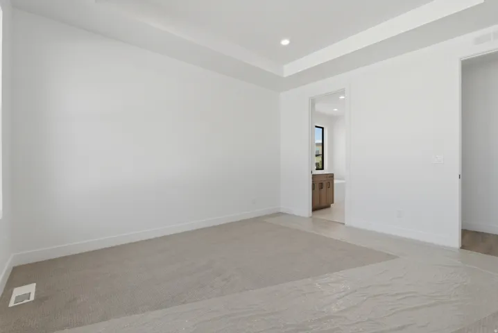 Spacious room featuring a tray ceiling with recessed lighting, light-colored carpeting, and a doorway leading to a bathroom with a wood-finish vanity