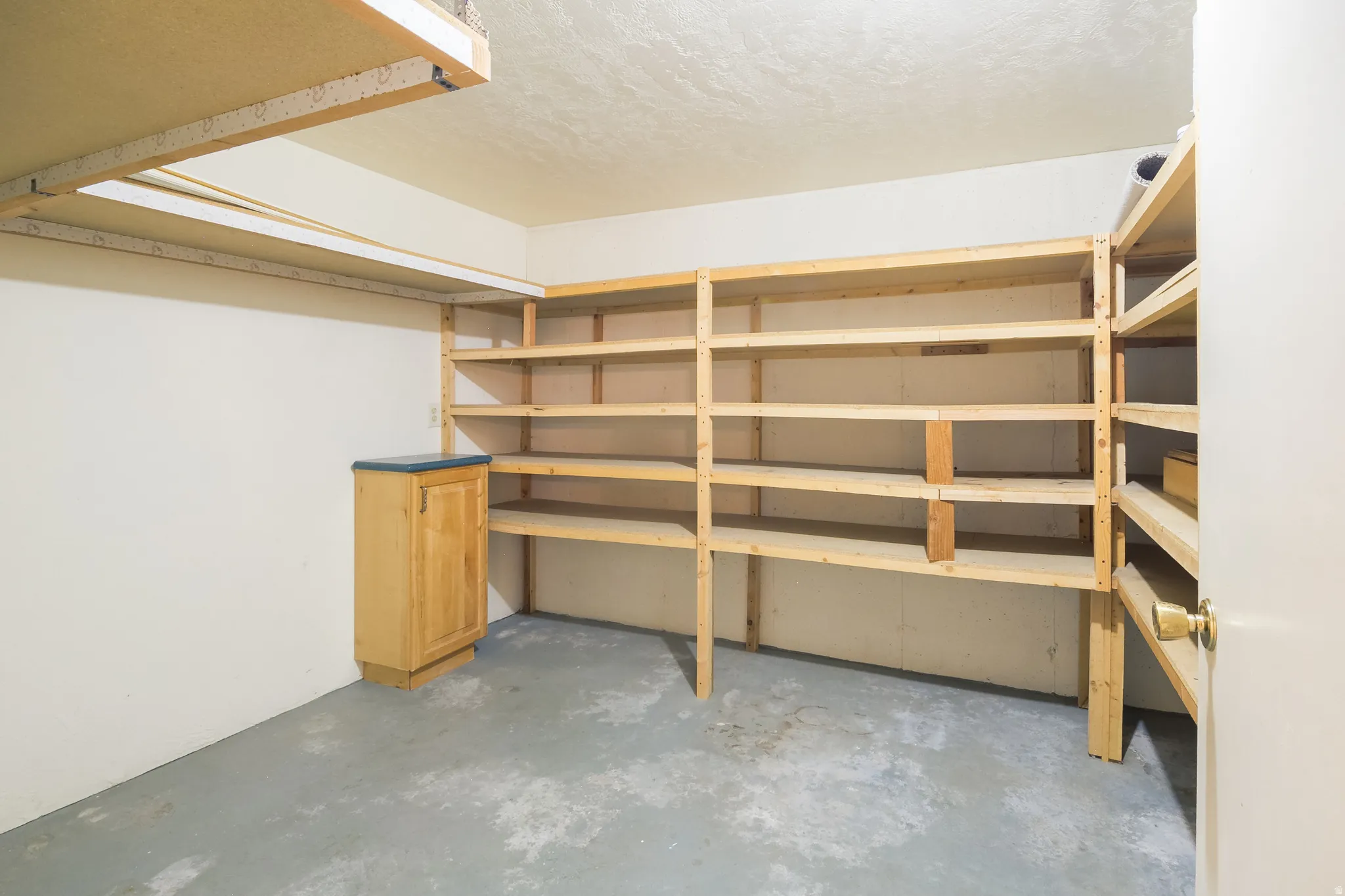 Ample storage room featuring extensive built-in wooden shelving, a light wood-finish base cabinet with a dark countertop, and durable concrete flooring