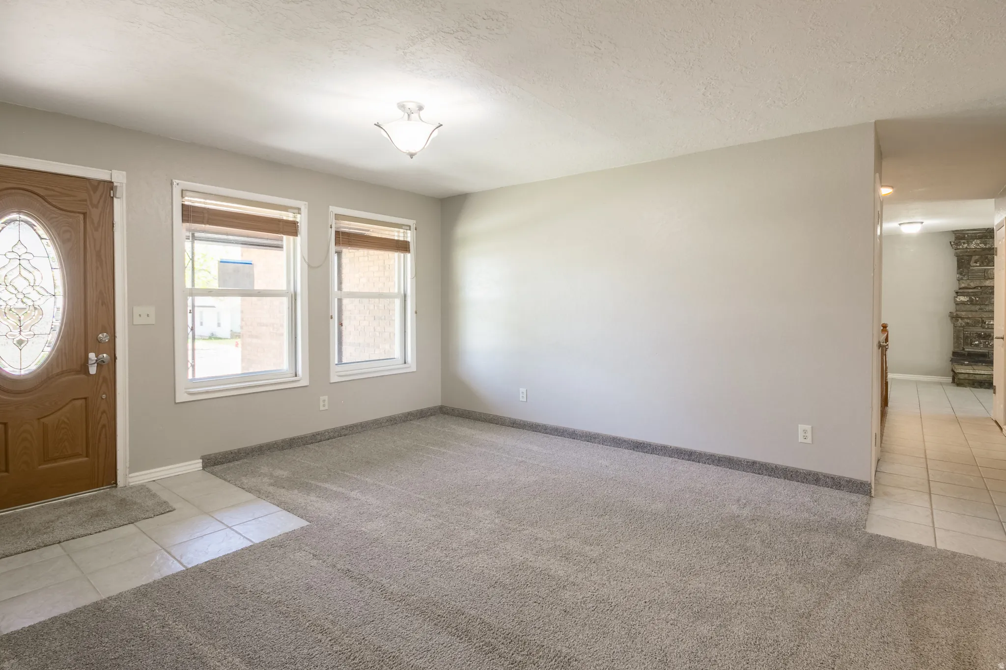 Entry space featuring a wood-finish door with an oval leaded glass insert, two windows with blinds, a ceiling-mounted light fixture, and textured gray carpet with a tiled entryway