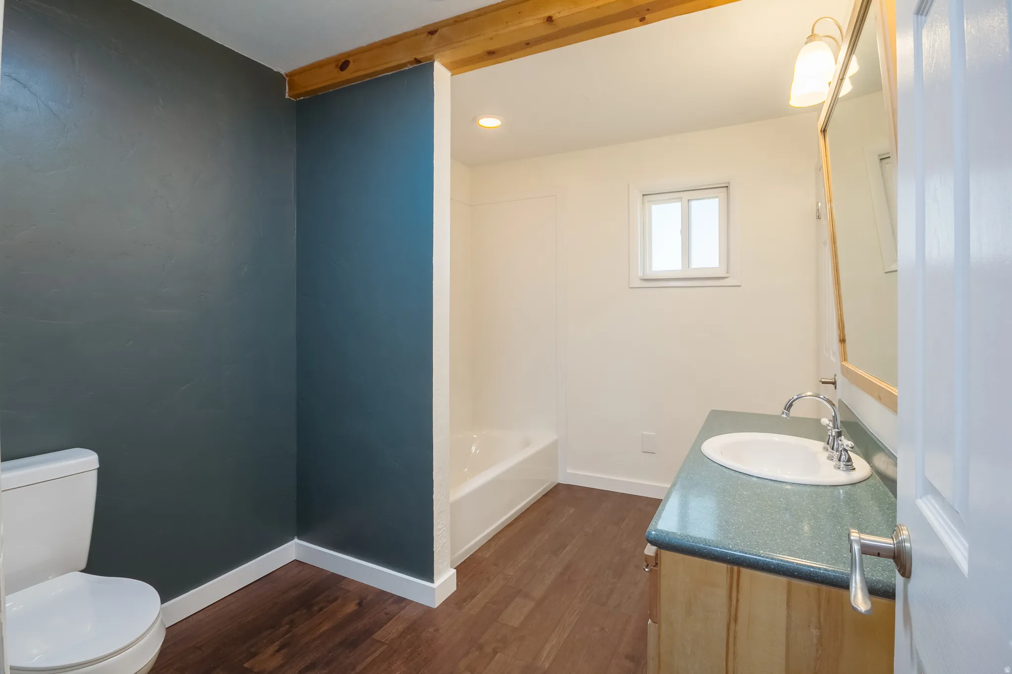Bathroom featuring wood-finish flooring, a white toilet, a built-in bathtub with shower, a vanity with an integrated sink, and a framed mirror