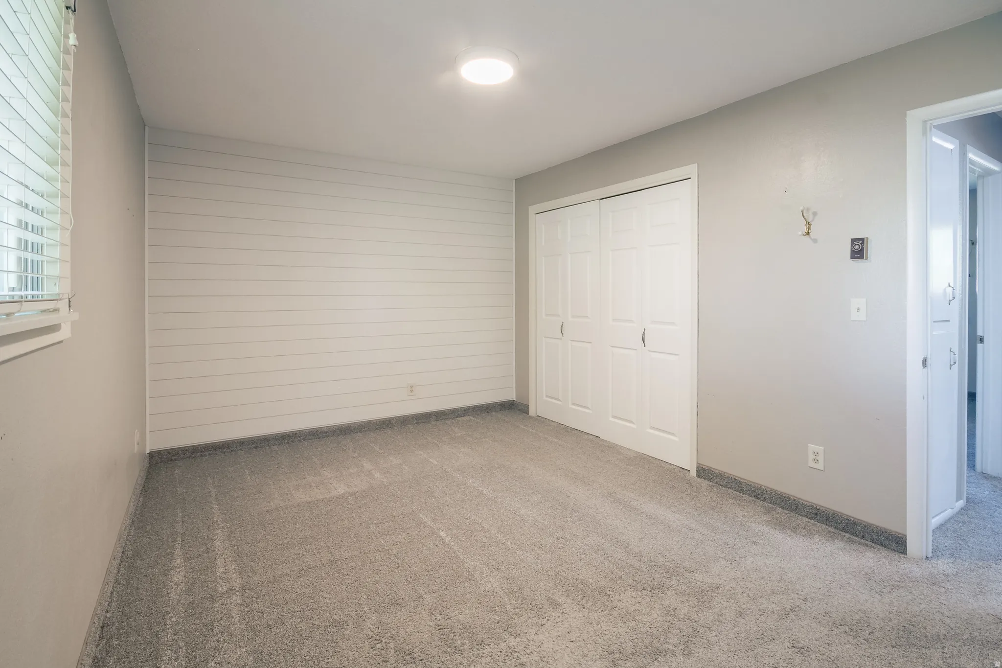 Room featuring light gray carpeting, a shiplap accent wall, white window blinds, and a double-door closet with white paneled doors