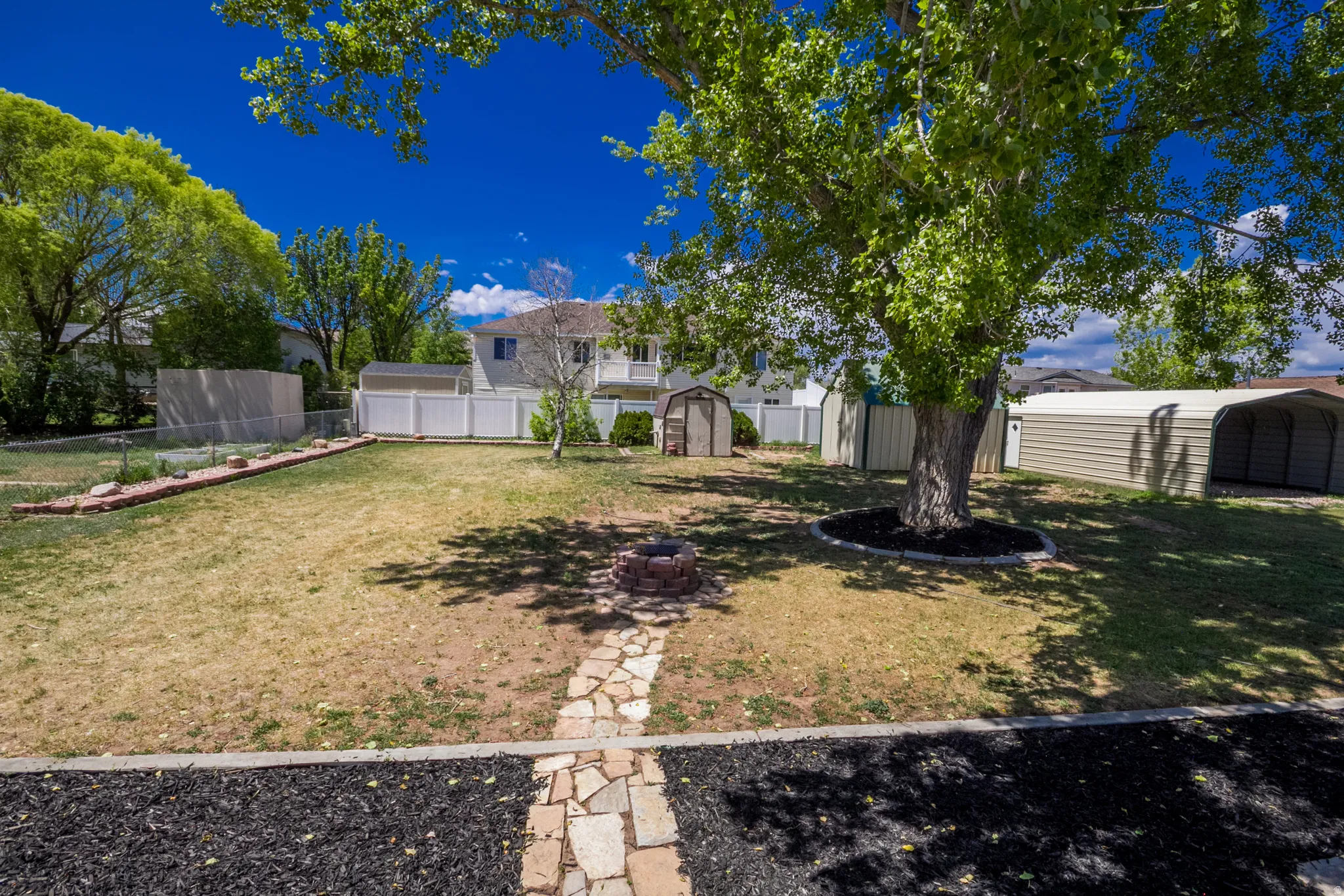 Expansive backyard featuring a mature tree with mulched base, a stone fire pit, and a flagstone pathway
