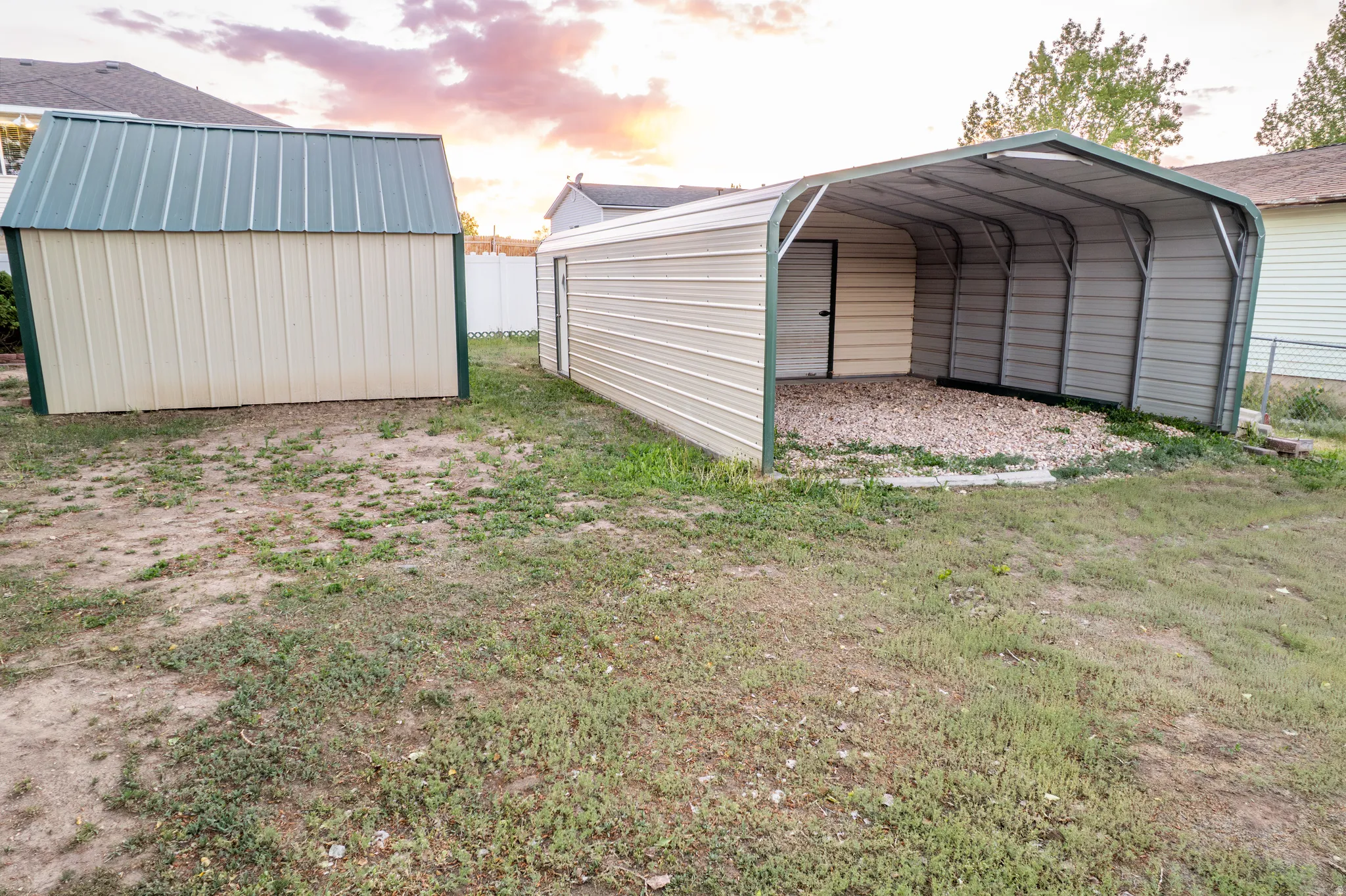 Expansive backyard featuring two metal storage structures: one fully enclosed with a green roof, and one open-sided carport with a gravel base and partial enclosure