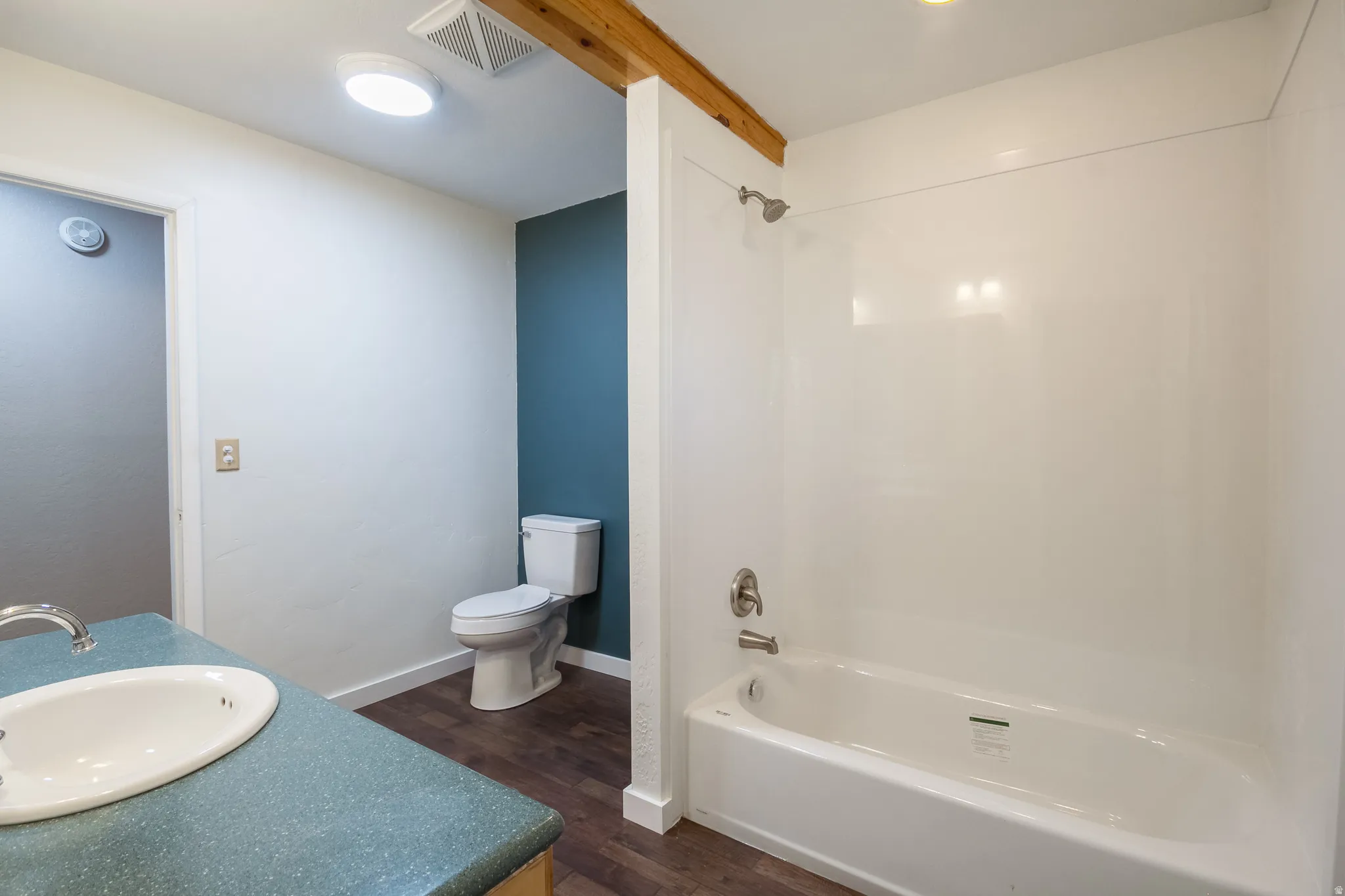 Bathroom featuring a white bathtub with a shower kit, a toilet, a vanity with an integrated sink and light blue countertop, and wood-finish flooring
