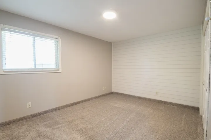 Room featuring light gray walls, plush gray carpeting, a single window with blinds, and a white shiplap accent wall