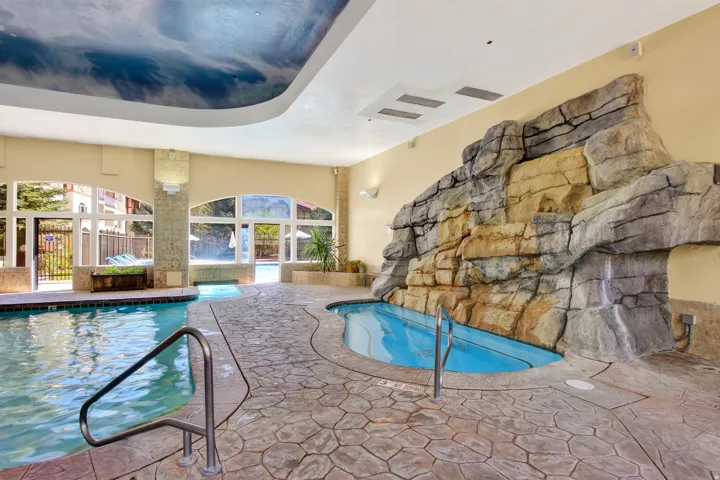 Indoor pool and spa area featuring a rock waterfall, flagstone-patterned flooring, and a cloud-mural ceiling