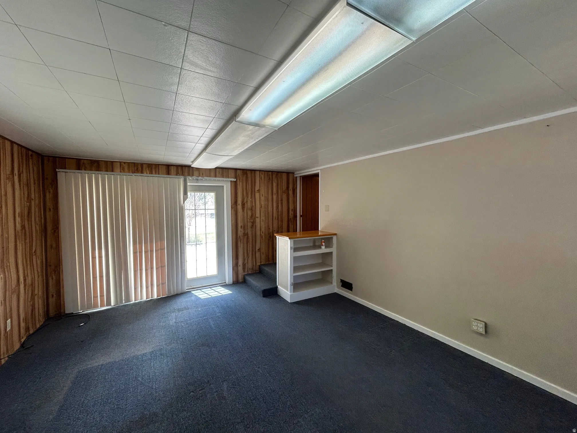 Room featuring blue carpet flooring, a drop ceiling, and a full-light door with vertical blinds