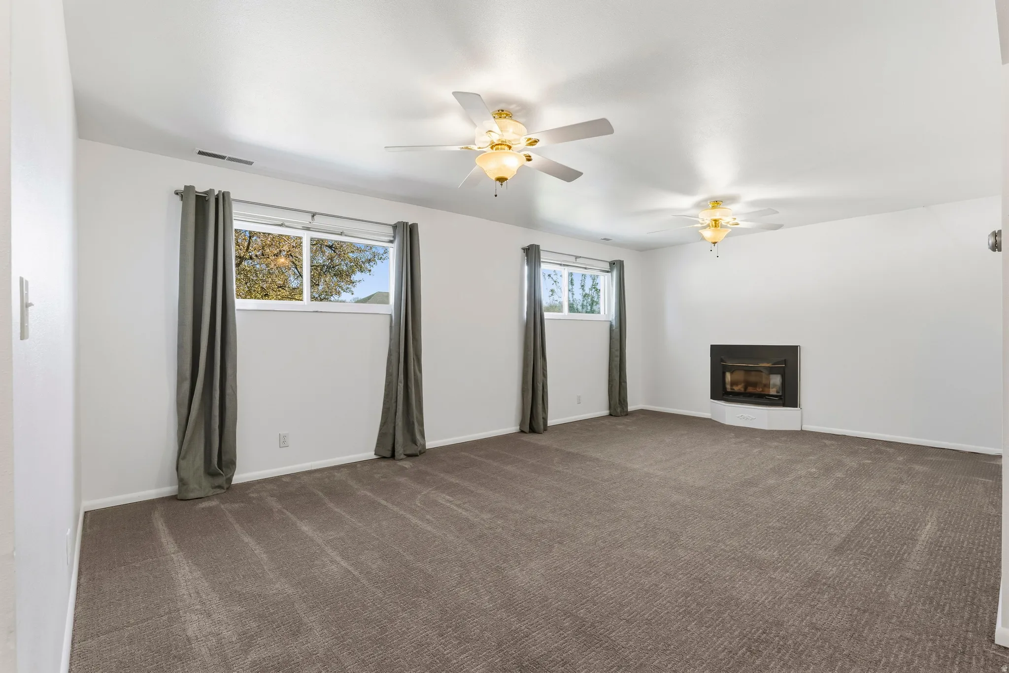 Spacious room featuring a corner-mounted fireplace with a dark surround, two windows, two ceiling fans with integrated lighting, and neutral-toned carpeting