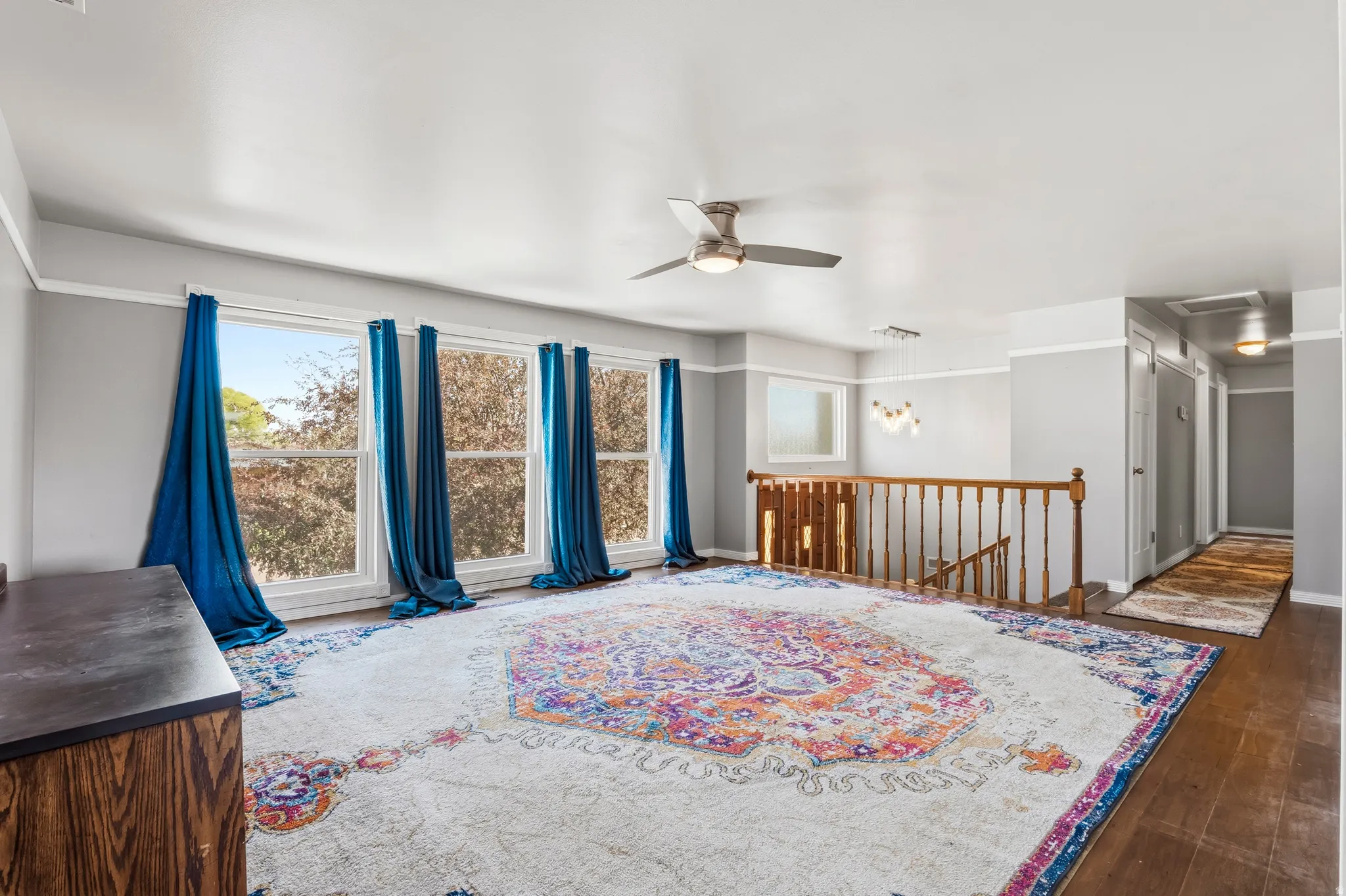 Spacious room featuring multiple large windows, a contemporary ceiling fan, rich wood-finish flooring, and a classic wooden staircase with railing