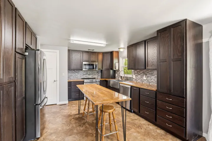 Spacious kitchen featuring dark wood cabinetry, stainless steel appliances, a mosaic tile backsplash, wood-finish countertops, and durable flooring