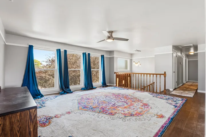 Spacious room featuring multiple large windows, a contemporary ceiling fan, rich wood-finish flooring, and a classic wooden staircase with railing