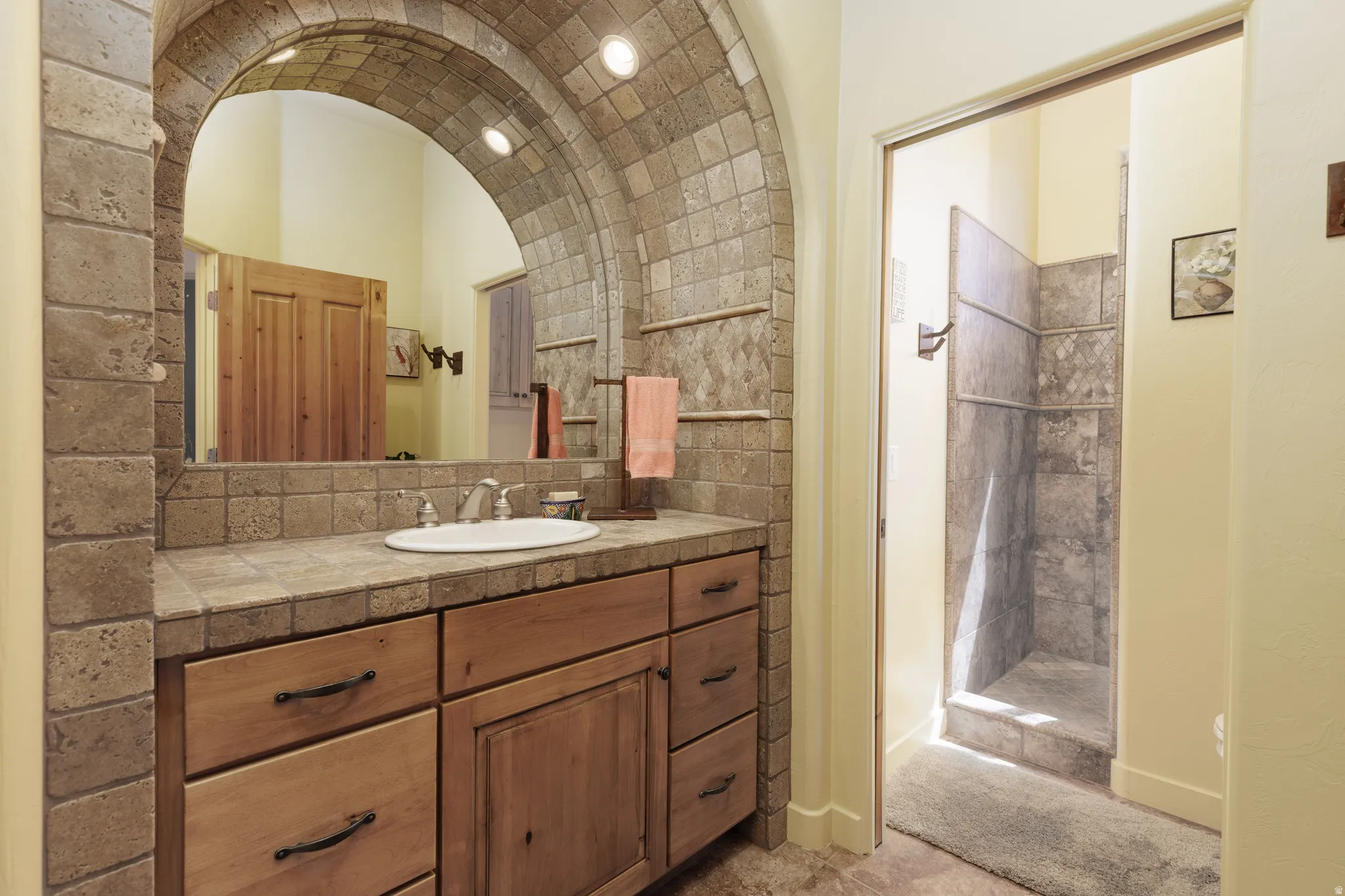 Bathroom featuring a wood vanity with a tile countertop, an undermount sink, and an arched stone-tiled mirror surround with recessed lighting