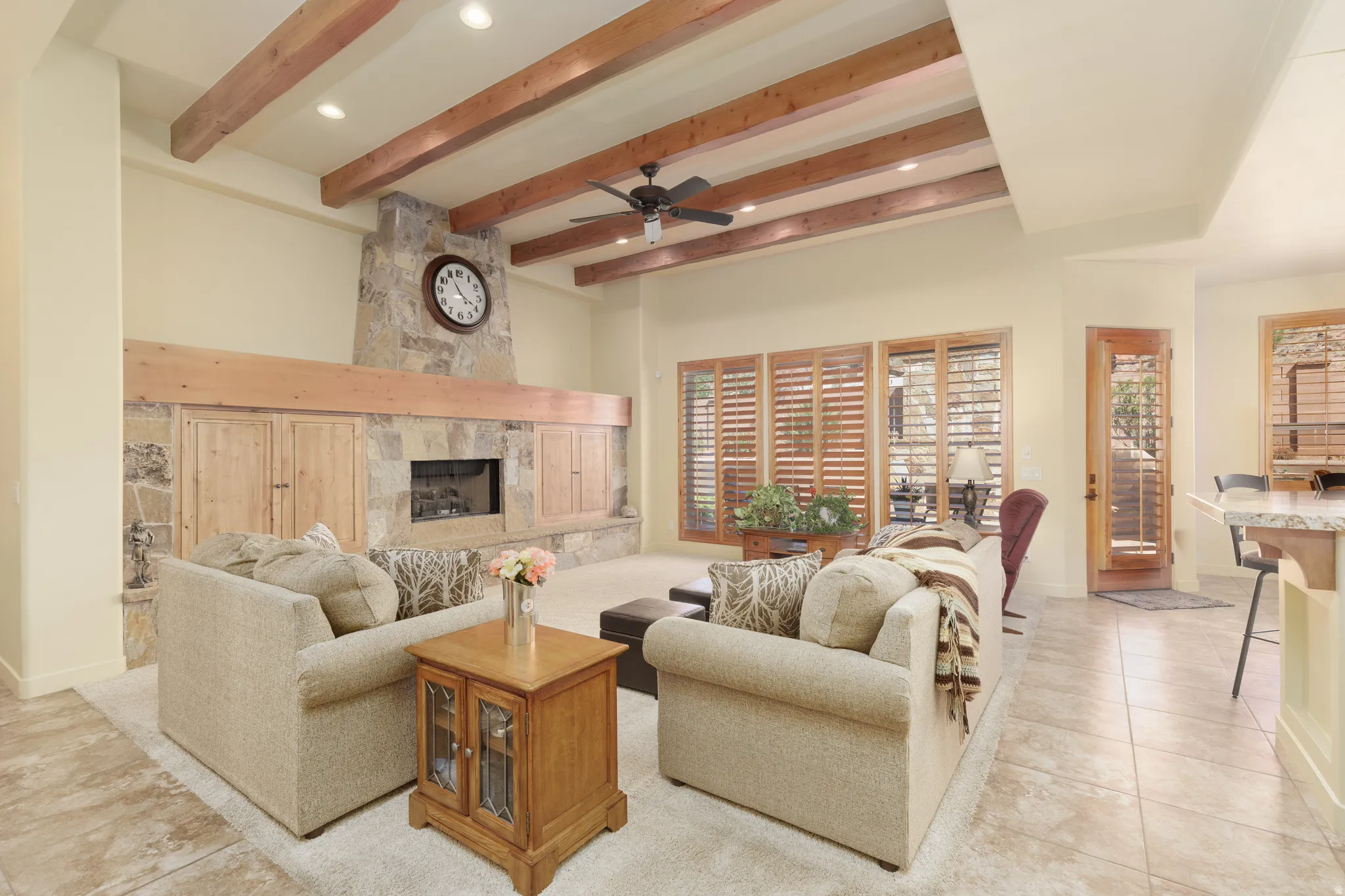 Spacious living area featuring a stone fireplace with a substantial wood mantel, integrated cabinetry, wood beam ceiling, recessed lighting, and wood-finish window shutters
