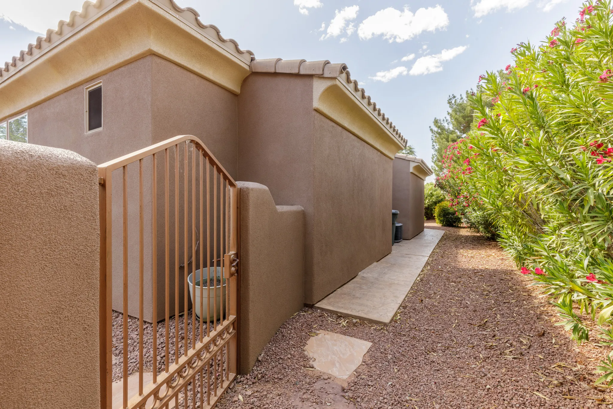 Stucco exterior featuring a decorative metal gate, tile roof accents, a concrete pathway, and a gravel-covered side yard with established green foliage