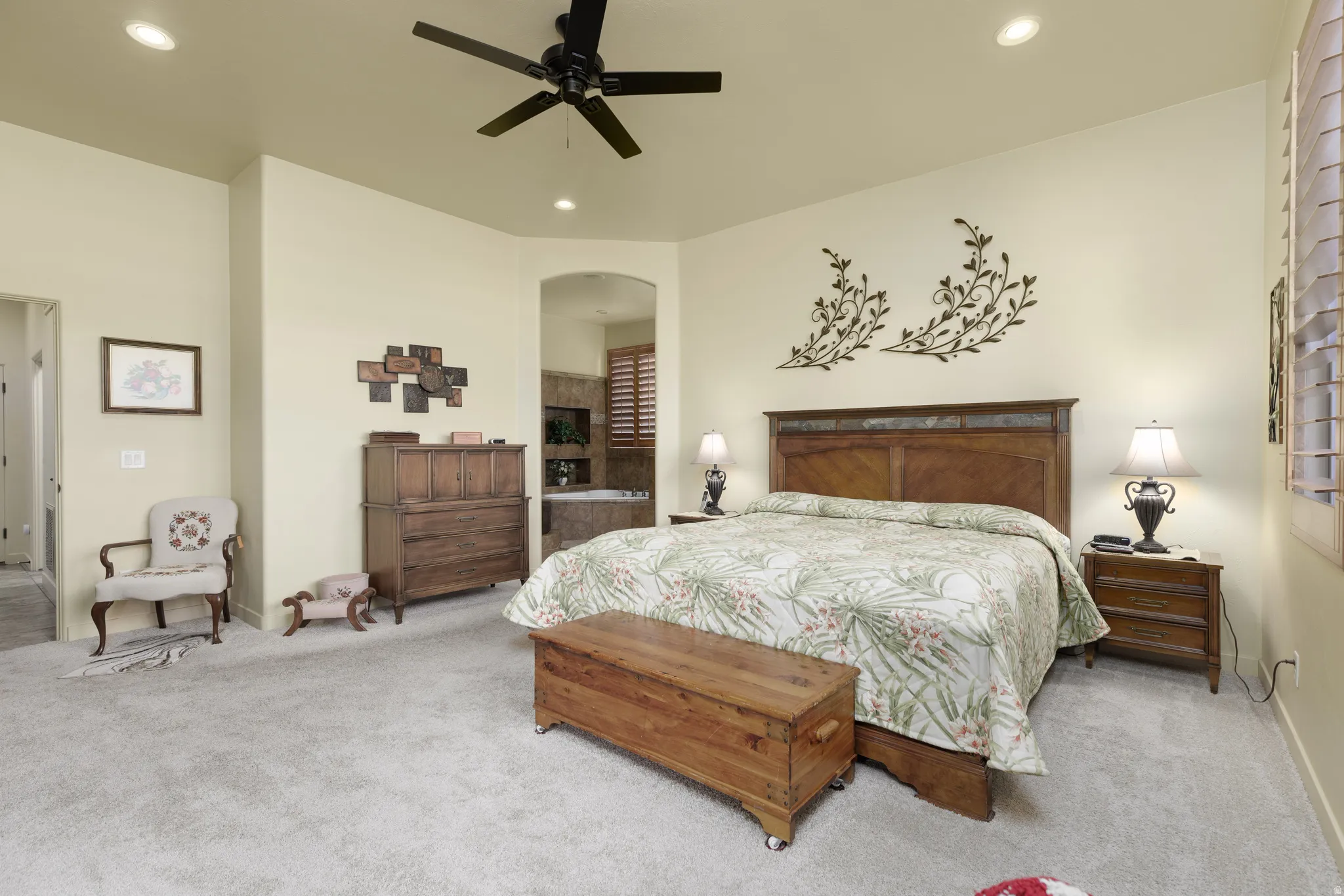 Carpeted interior space featuring a ceiling fan with integrated lighting, recessed ceiling lights, and an arched entry to an en-suite bathroom with a built-in tub and louvered window coverings