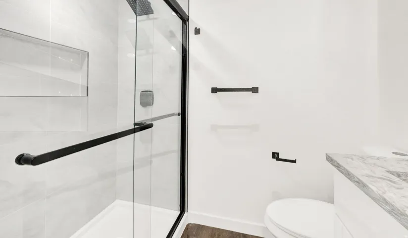 Contemporary bathroom featuring a frameless glass shower enclosure with matte black hardware, a recessed shower niche, light-toned wall tiling, and a vanity with a stone countertop