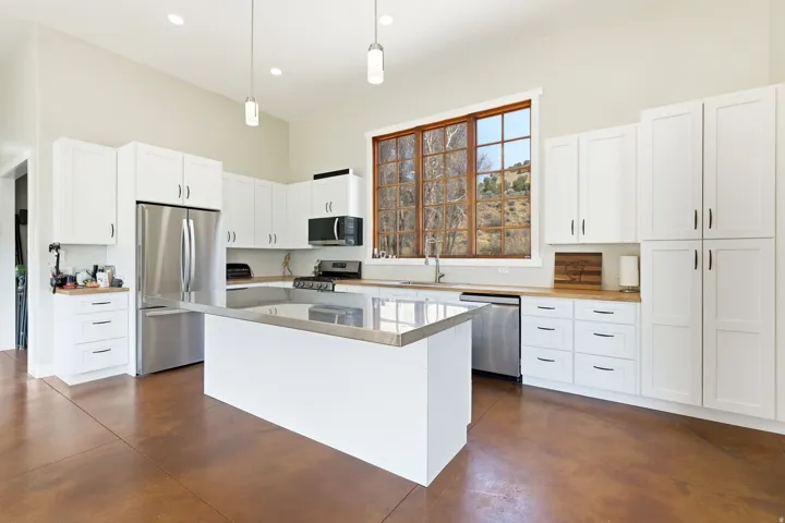 Spacious kitchen featuring an expansive island with a stainless steel countertop, white cabinetry with black hardware, stainless steel appliances, wood-finish countertops, and large windows
