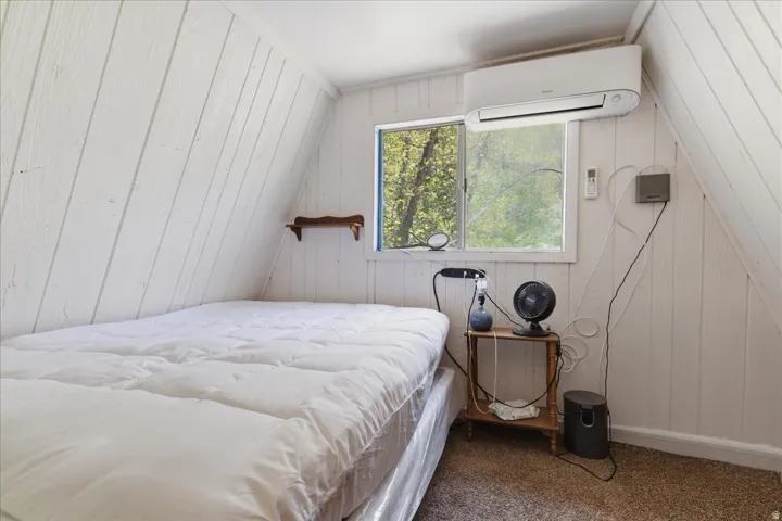 Slanted white paneling throughout, a wall-mounted mini-split unit, and a single pane window with a forest backdrop