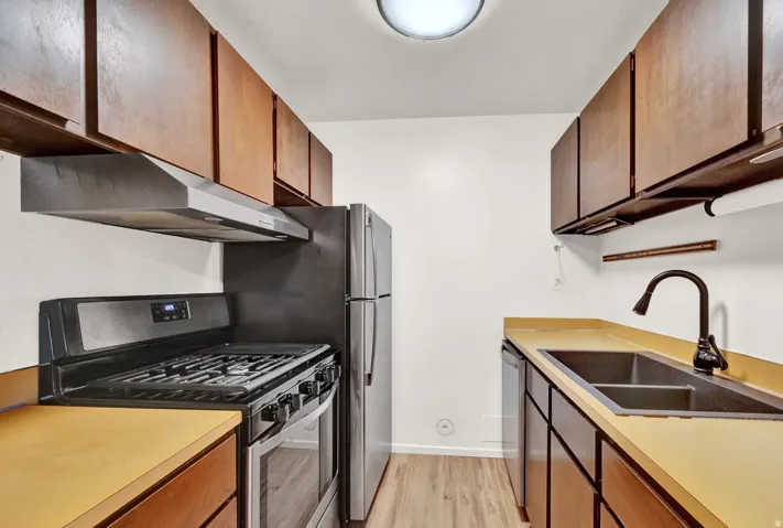 Galley kitchen featuring wood-finish cabinetry, light-toned countertops, a black gas range with an overhead vent hood, a stainless steel refrigerator, and a double basin sink with a dark-finish faucet