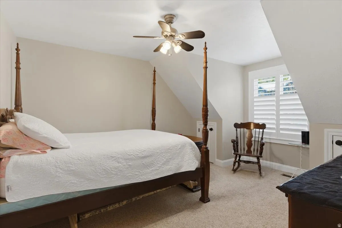 Carpeted room featuring a ceiling fan with integrated lighting, a window with white plantation shutters, and a sloped ceiling design