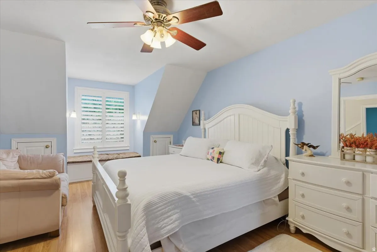 Bedroom featuring warm wood-finish flooring, a ceiling fan with integrated lighting, and a window with white plantation shutters