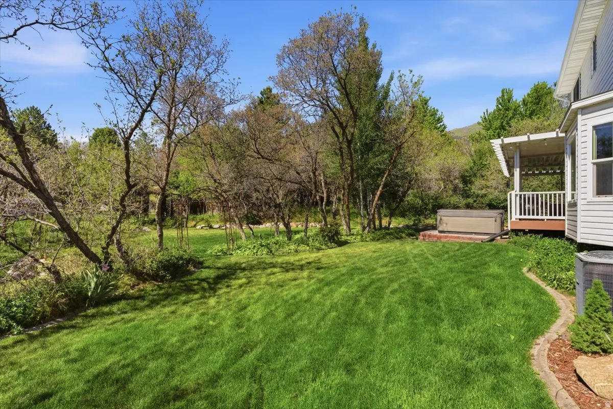 Expansive green lawn surrounded by mature trees, featuring a covered wooden deck with white railing, and a hot tub on a brick patio