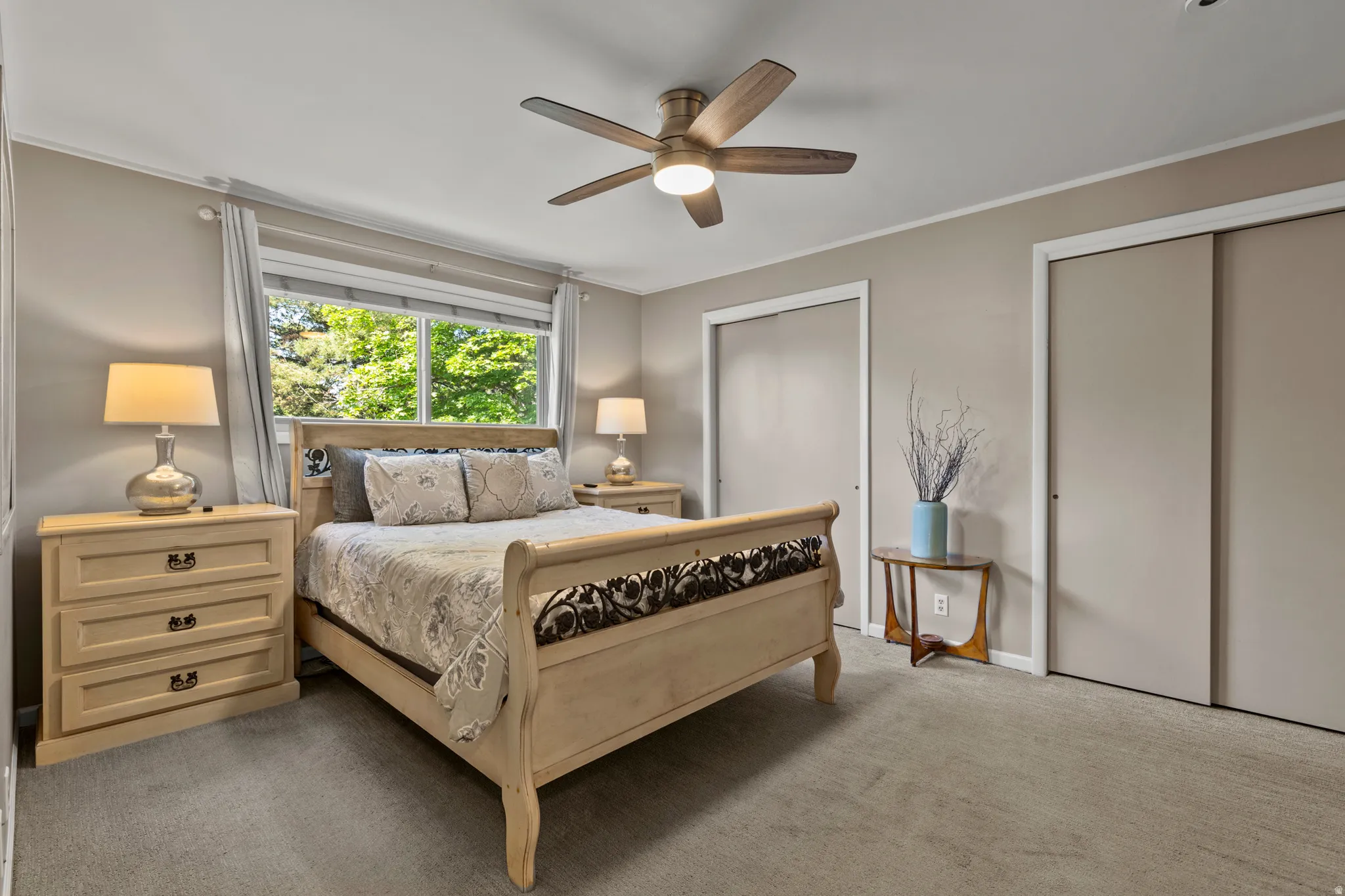Carpeted bedroom with a window overlooking green trees, featuring a ceiling fan with integrated lighting and two sliding door closets