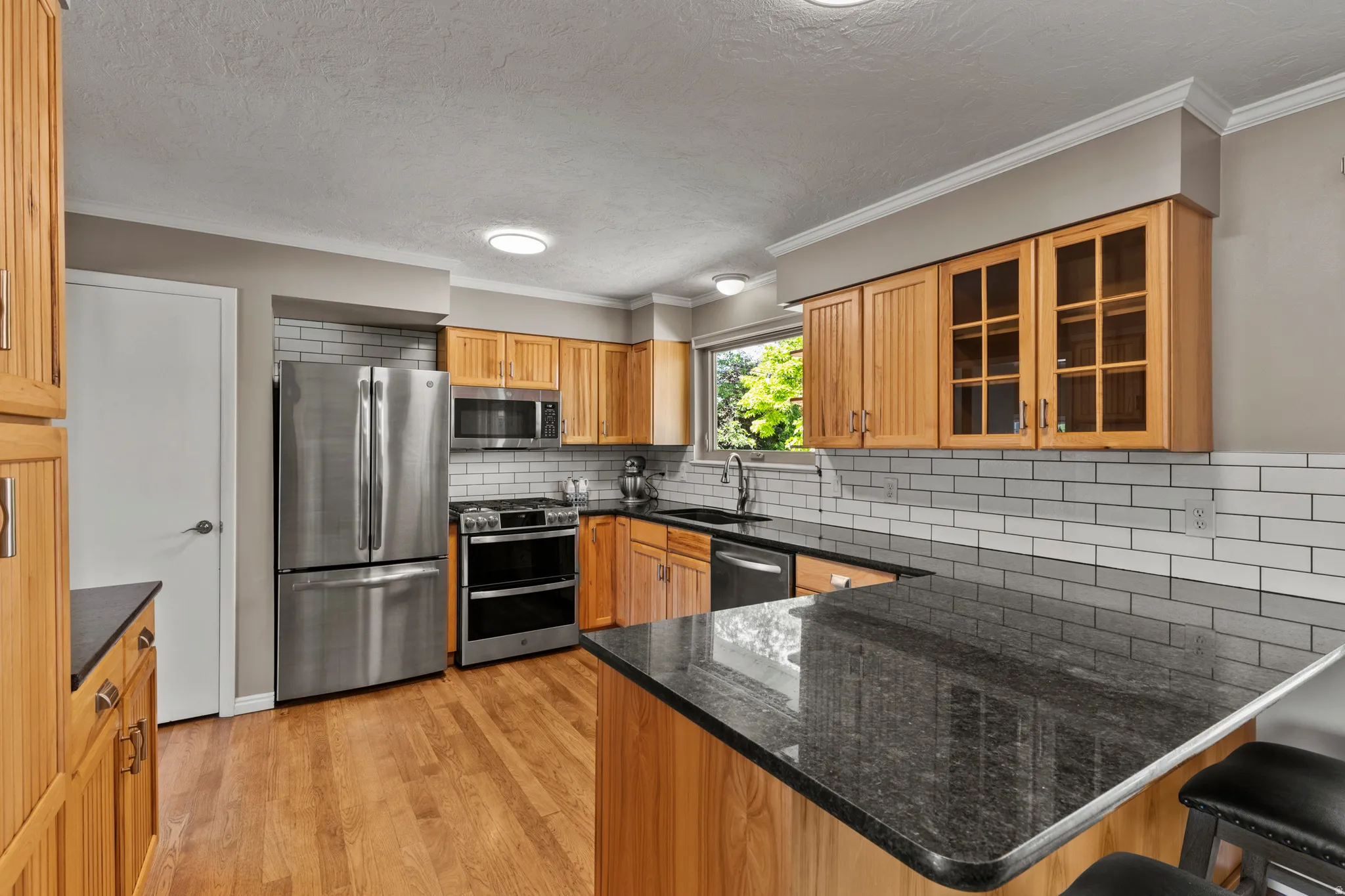 Kitchen featuring wood-finish flooring, stainless steel appliances, granite countertops, subway tile backsplash, and light wood cabinetry