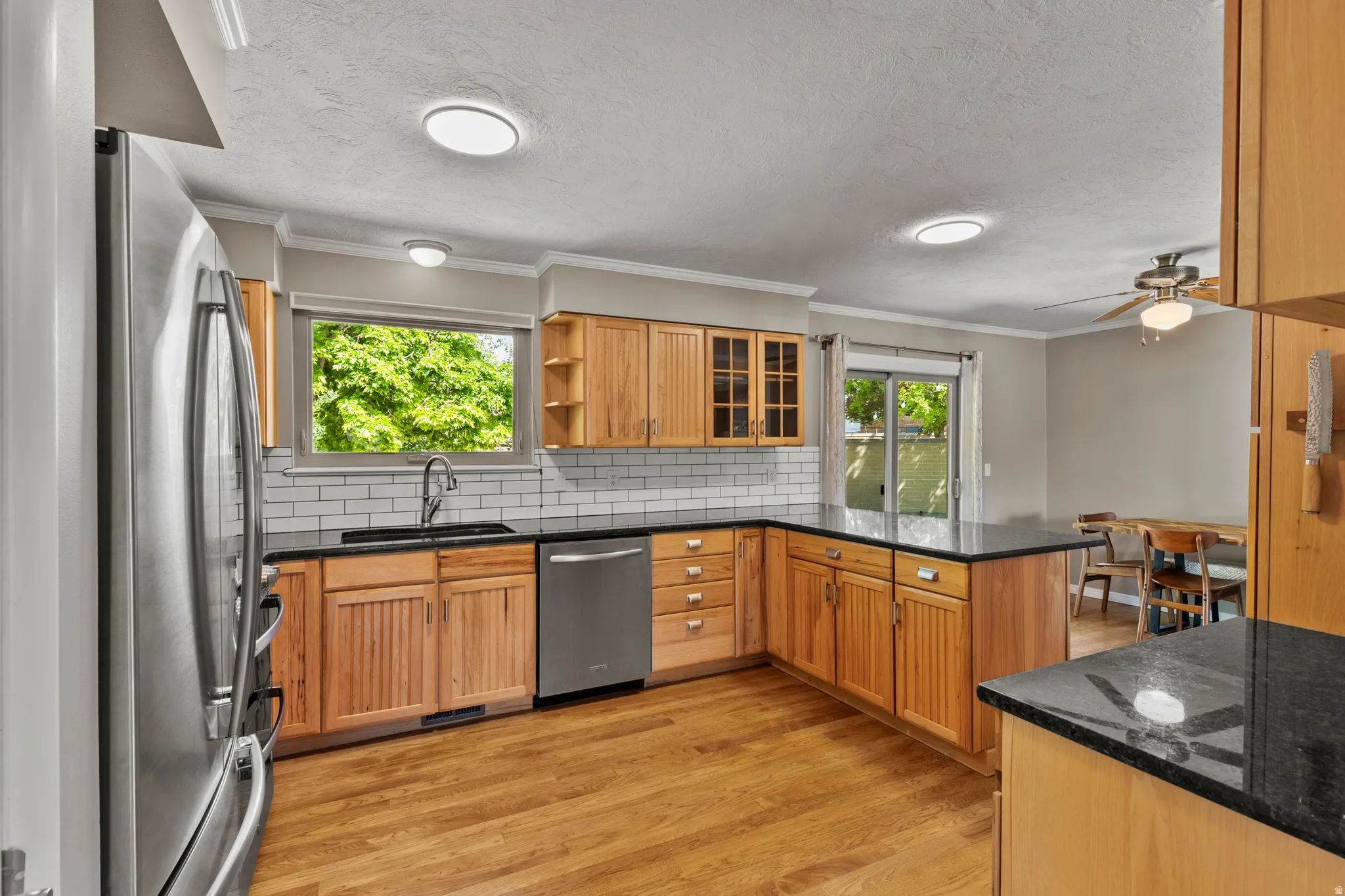 Kitchen featuring wood cabinetry, stainless steel appliances, black countertops, a subway tile backsplash, and wood-finish flooring