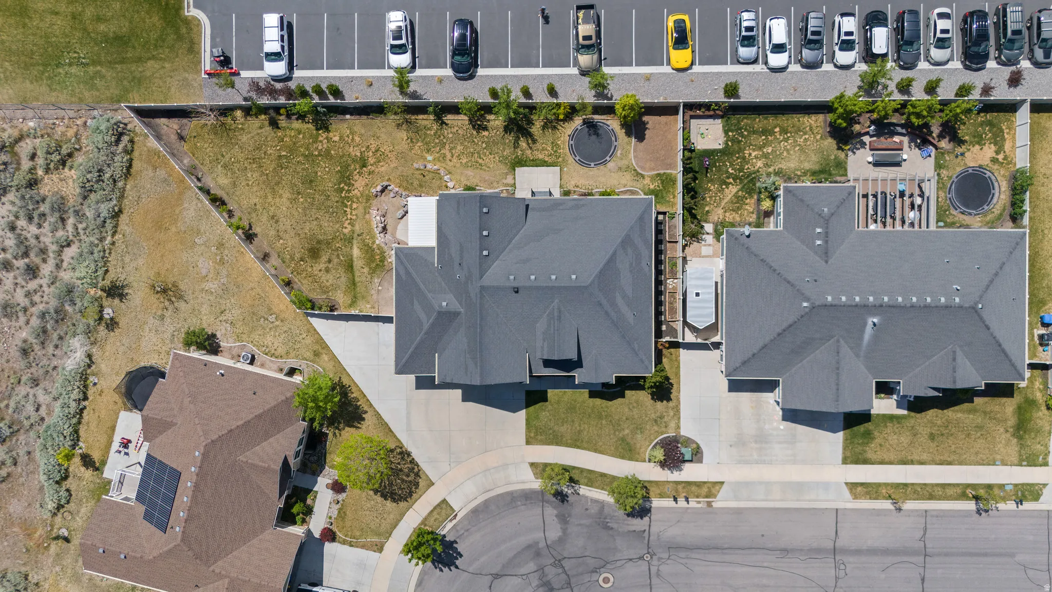 Aerial perspective showcasing a property with a dark shingle roof, concrete driveway, and surrounding green landscaping