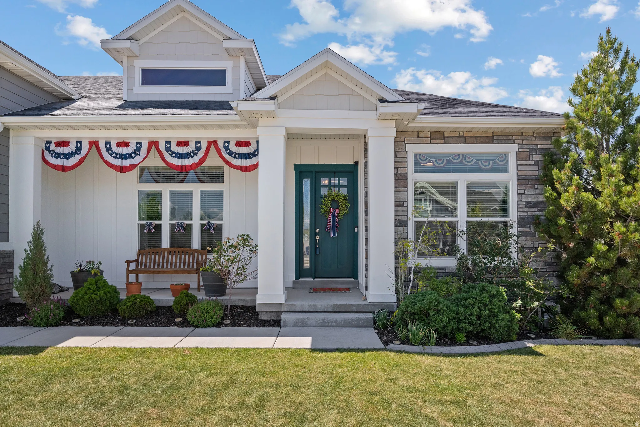Contemporary home exterior featuring a dark teal entry door, stone and board and batten siding, multiple windows, a covered porch, and mature landscaping