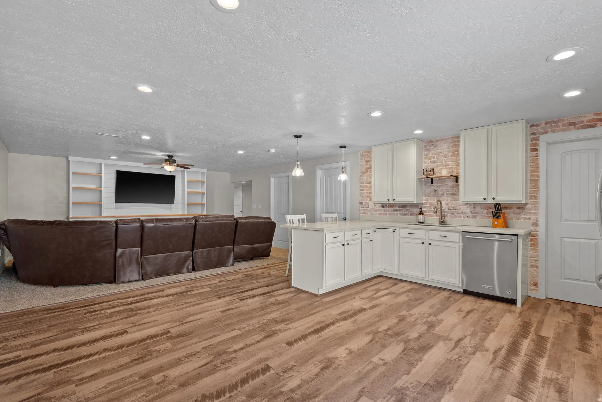 Open-concept living space featuring wood-finish flooring, a contemporary kitchen with white cabinetry and a brick backsplash, recessed lighting, and a built-in media center with shelving
