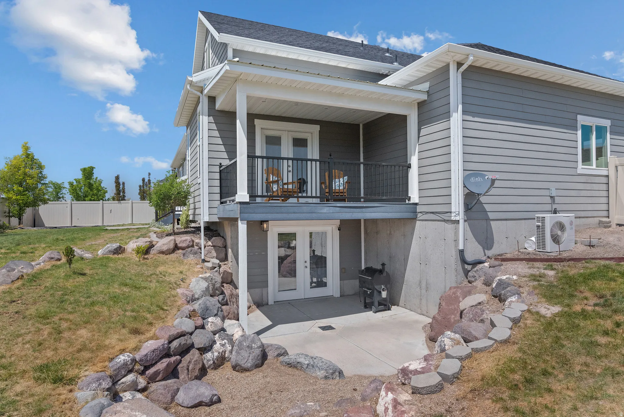 Gray siding exterior with a covered upper-level balcony, ground-level patio, rock landscaping, and a fenced perimeter