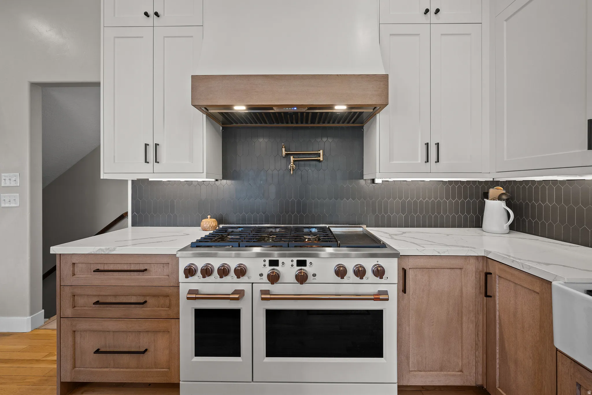 Gourmet kitchen featuring a professional-grade range, white shaker-style upper cabinetry, wood-finish lower cabinetry, white stone countertops, and a dark picket tile backsplash