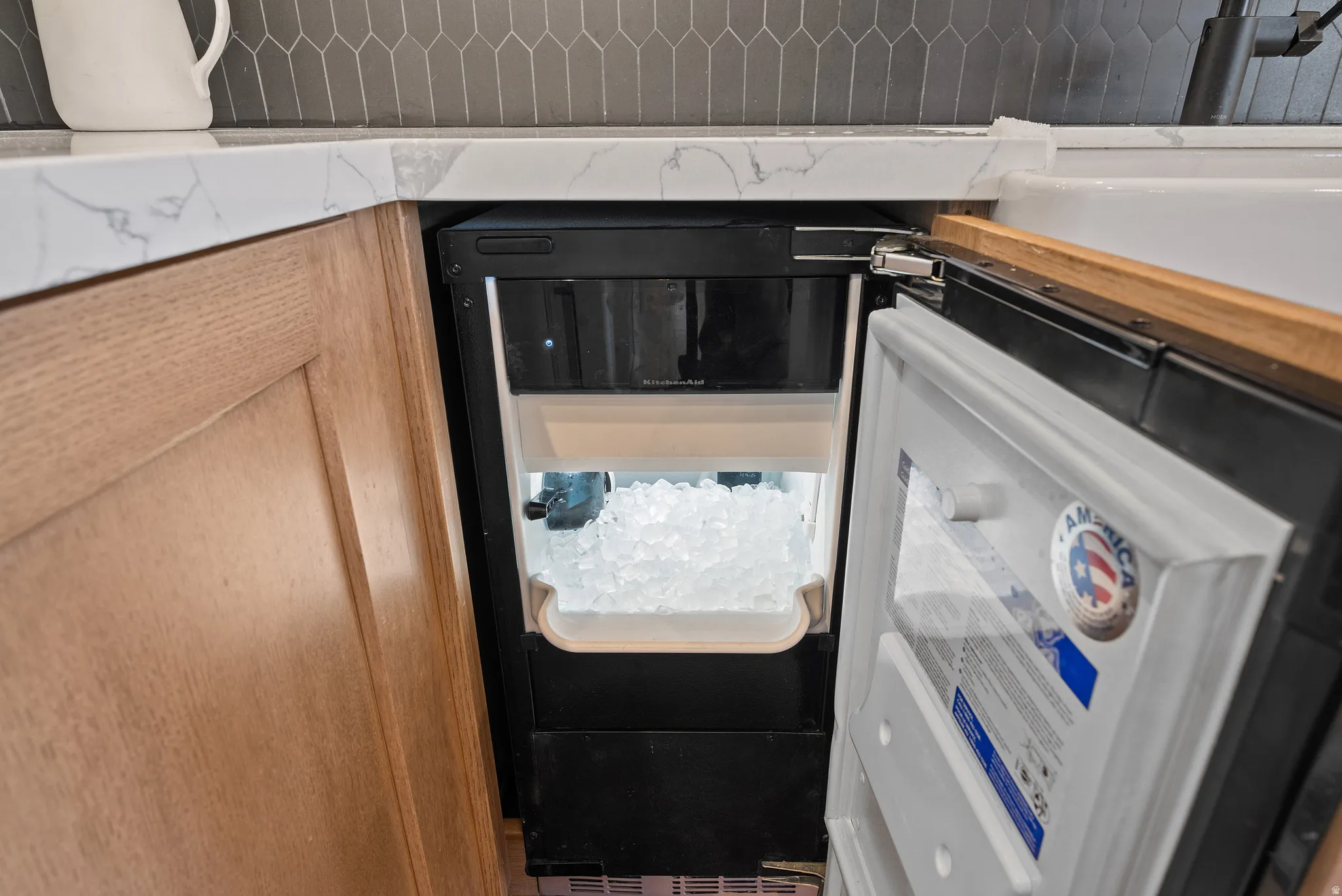 Built-in under-counter ice maker with a full bin of ice, set beneath a white countertop with grey veining
