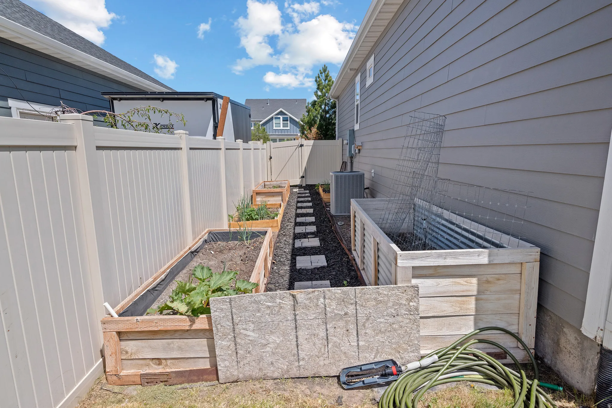 Side yard featuring a series of wooden raised garden beds, a light-toned vinyl privacy fence, and a stone paver pathway