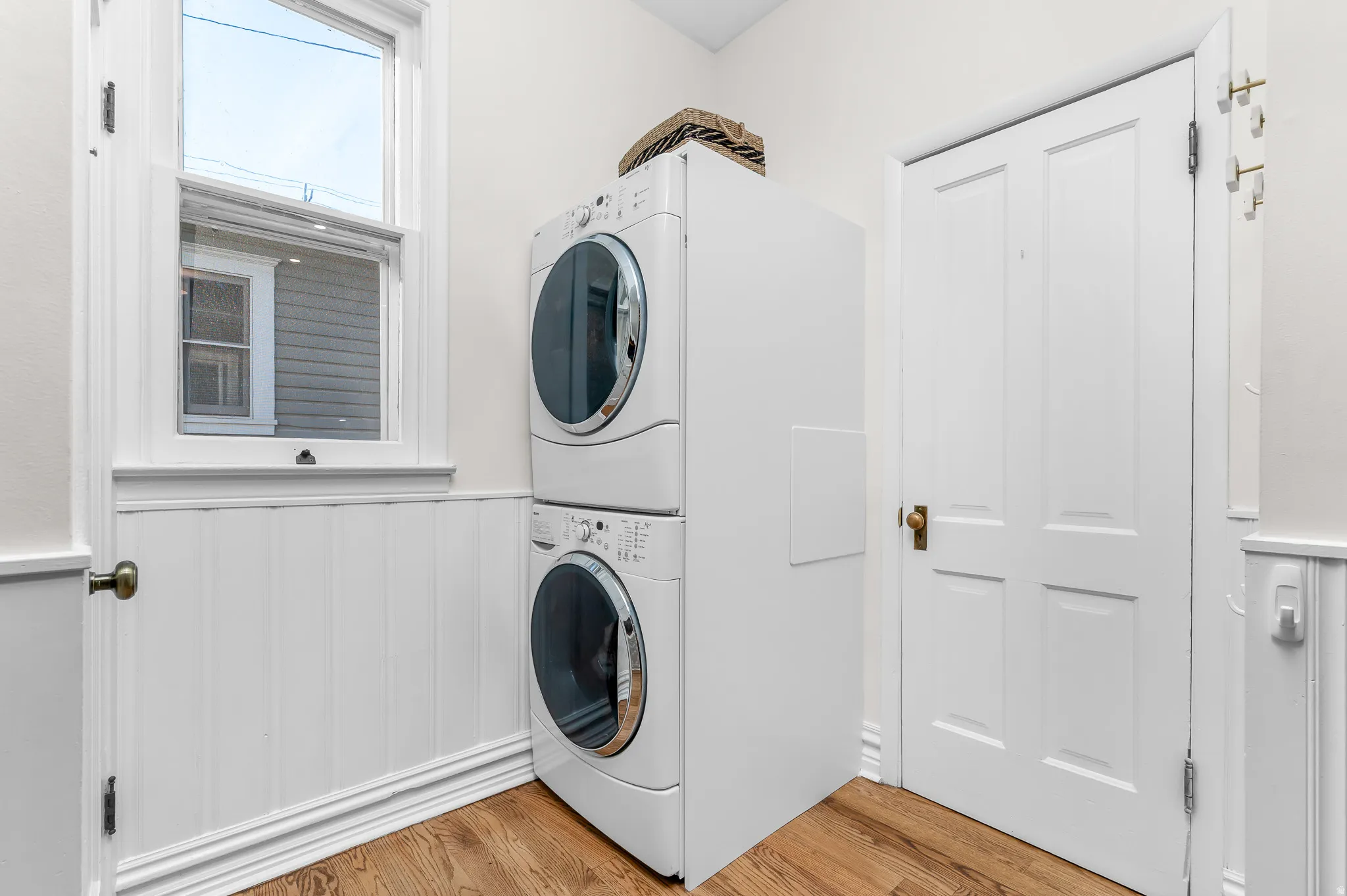 Laundry area featuring a stacked washer and dryer, wood-finish flooring, white paneled walls, a single-pane window, and a four-panel door with brass hardware