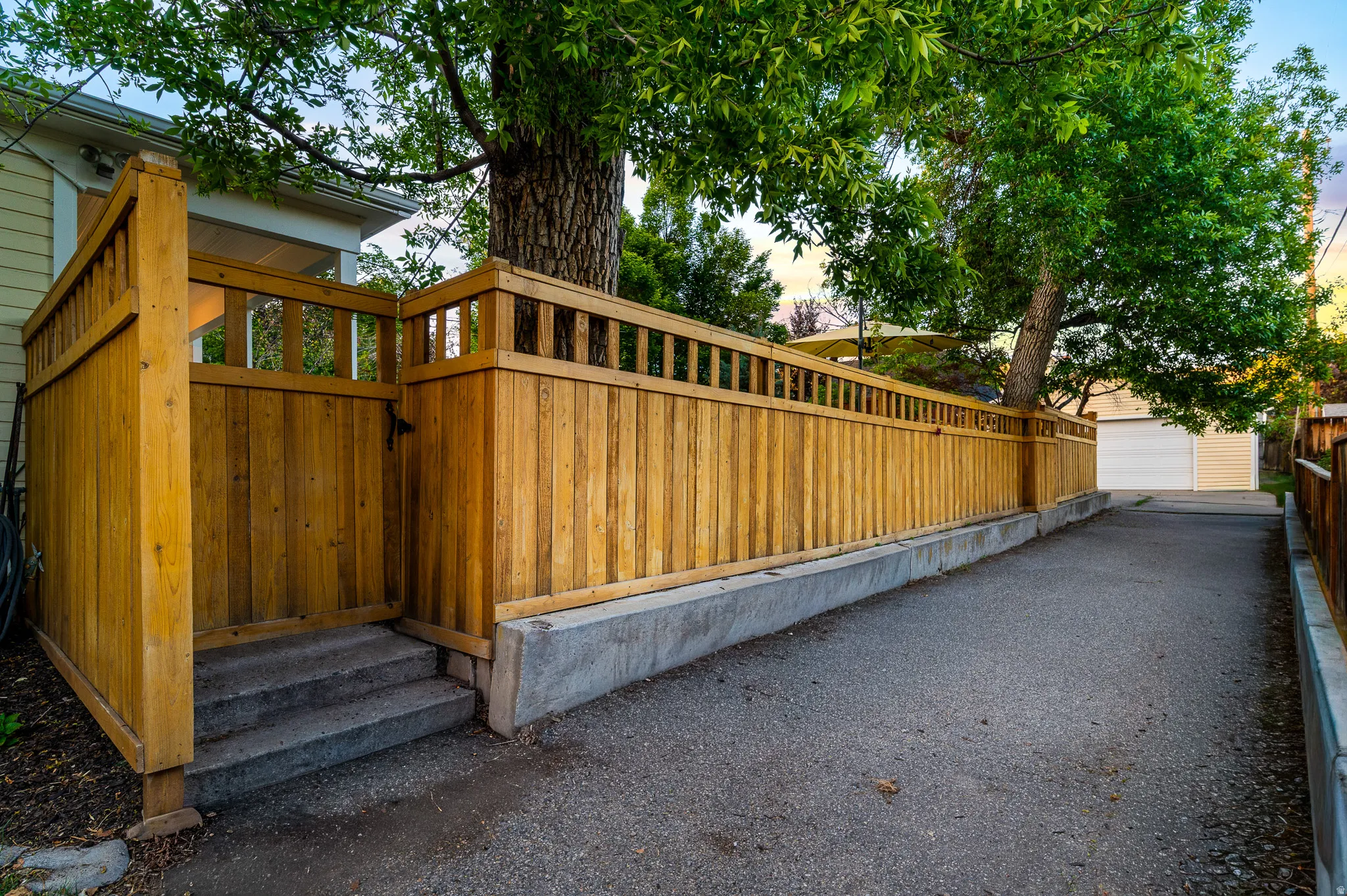 Paved driveway bordered by a wood privacy fence on a concrete base, featuring a gated entrance with steps and mature trees