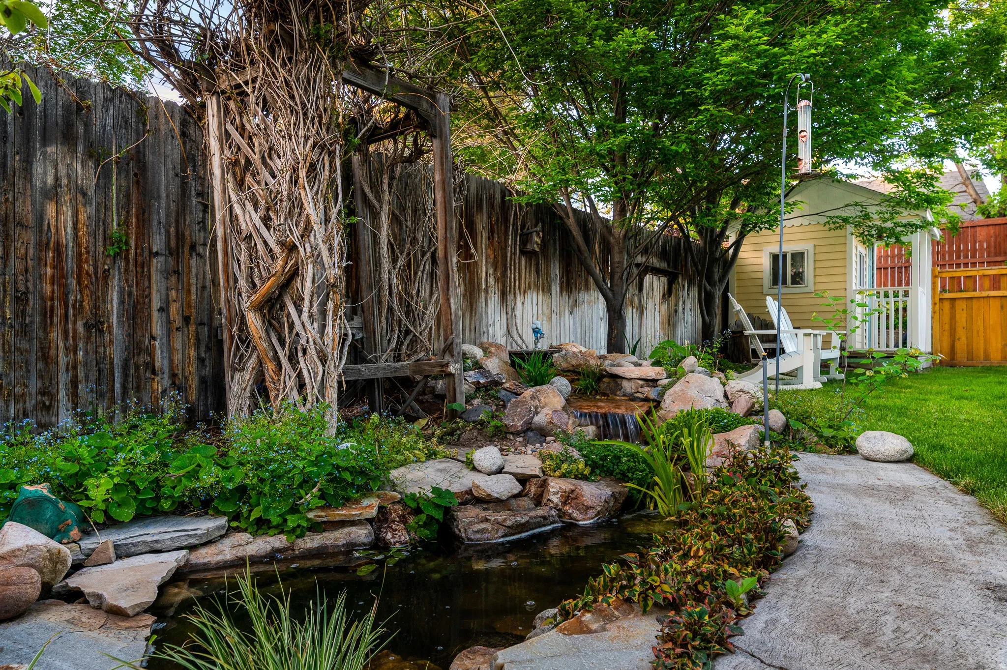 Lush yard featuring a rock waterfall, pond, concrete pathway, and mature trees