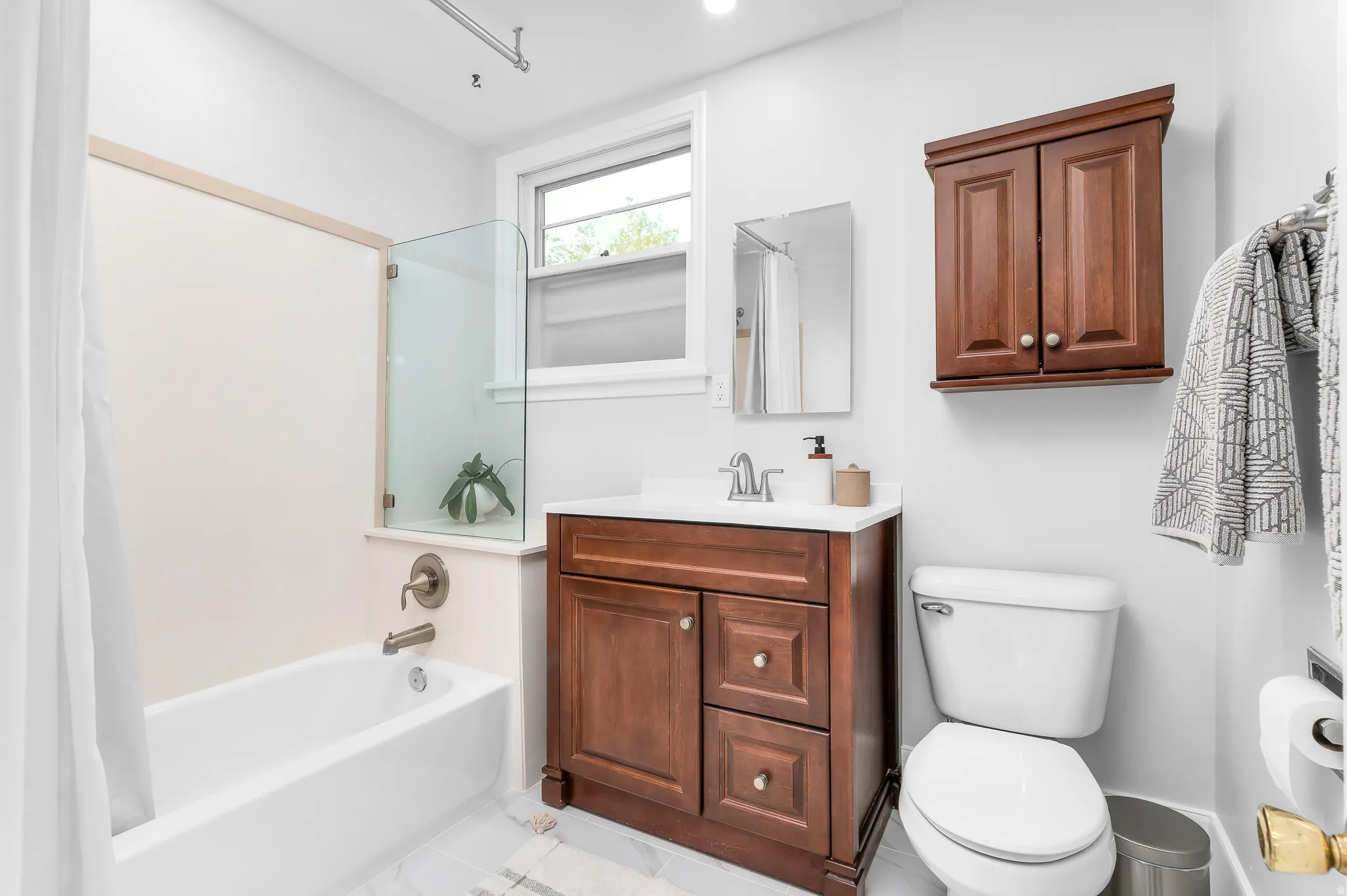 Bathroom featuring a built-in bathtub with a glass partition, wood-finish vanity with countertop sink, wall-mounted mirror, and a window providing natural light