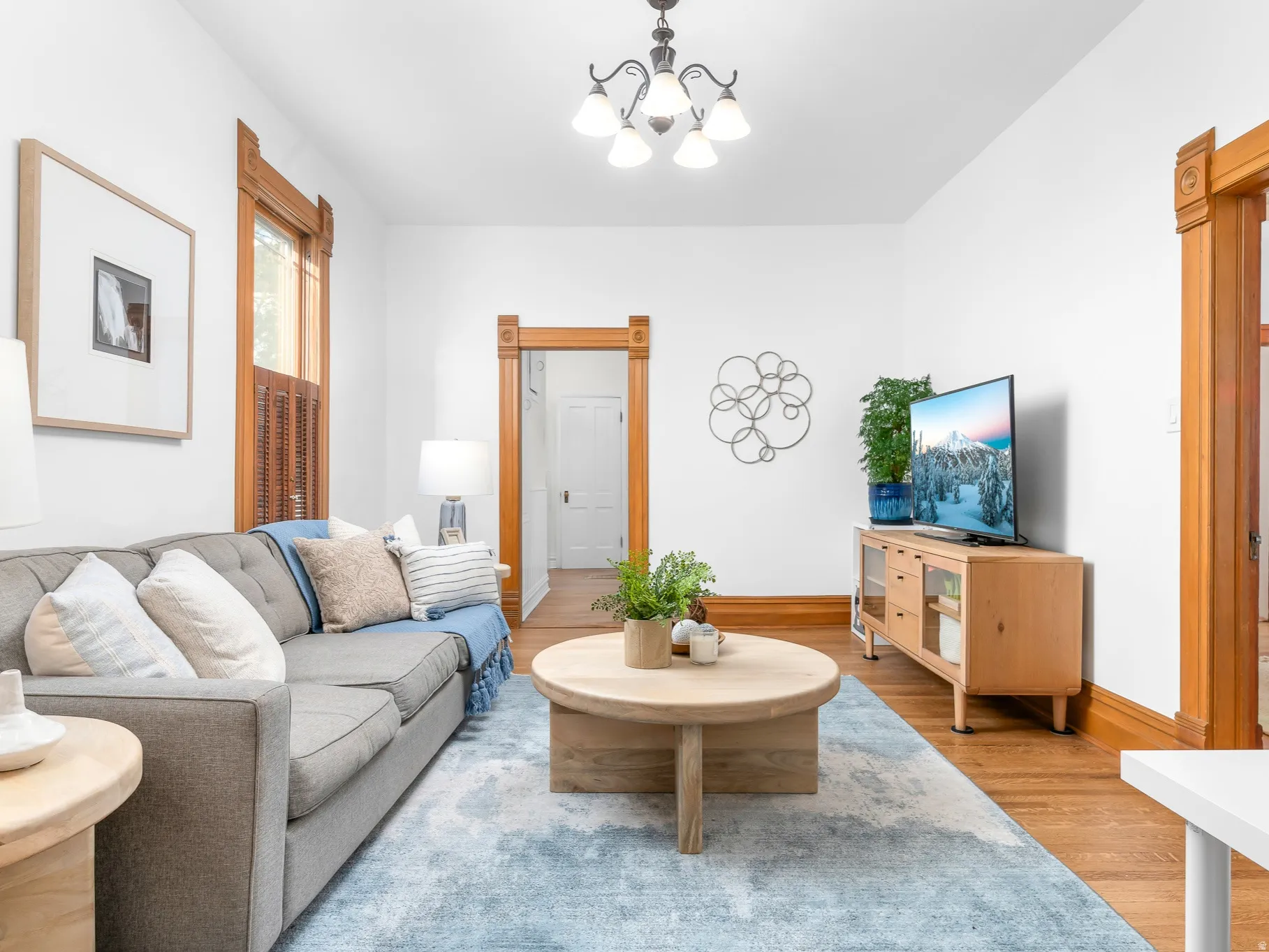 Living area featuring white walls, wood-finish flooring, a decorative ceiling light fixture, and ornate wood trim around windows and doorways