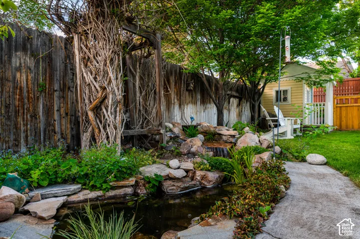 Lush yard featuring a rock waterfall, pond, concrete pathway, and mature trees