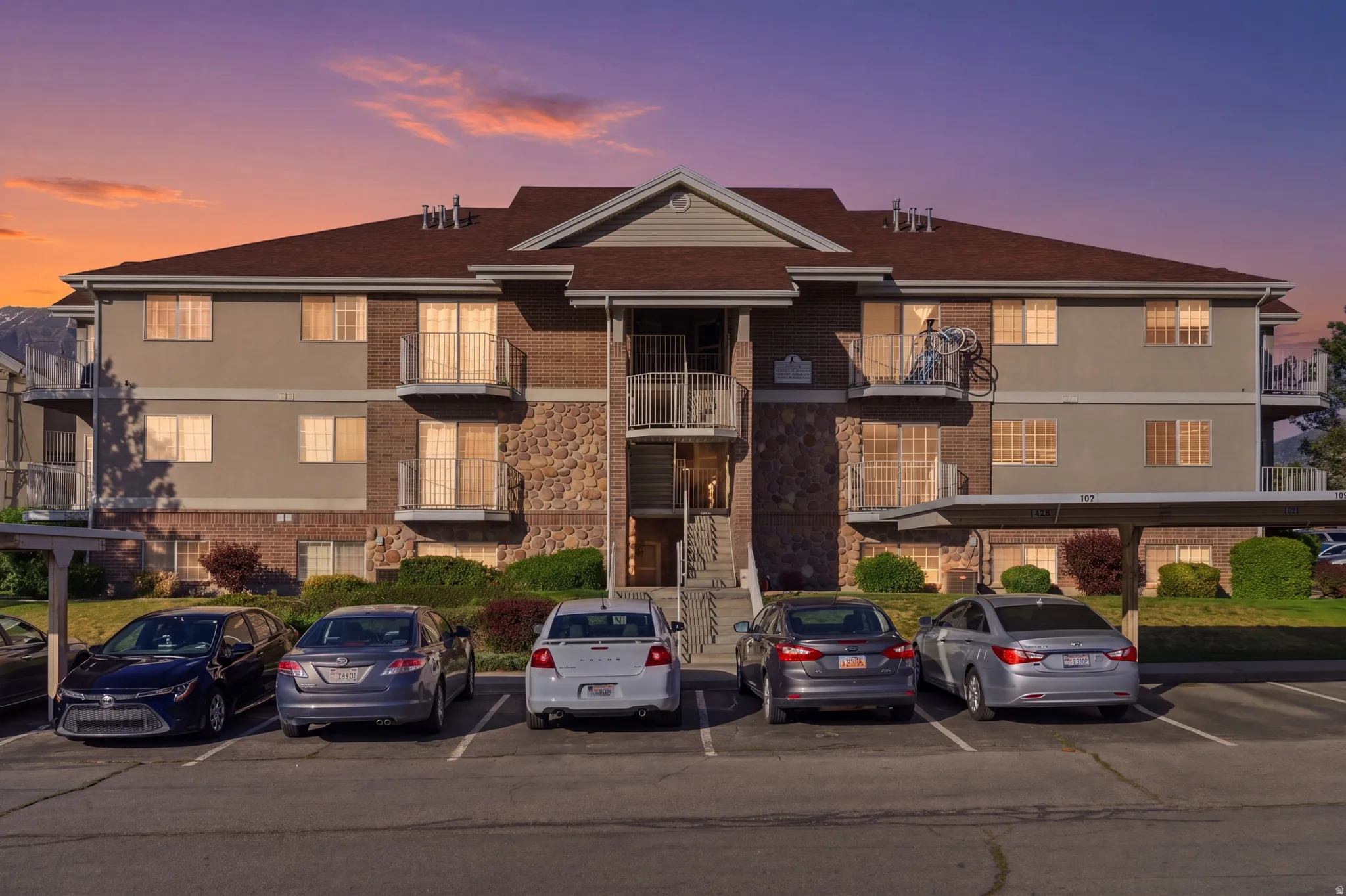 Multi-story building featuring a brown shingle roof, stone and stucco facade, private balconies, and covered parking spaces